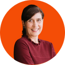 A woman with short dark hair, smiling, wearing a maroon sweater, standing against an orange background.