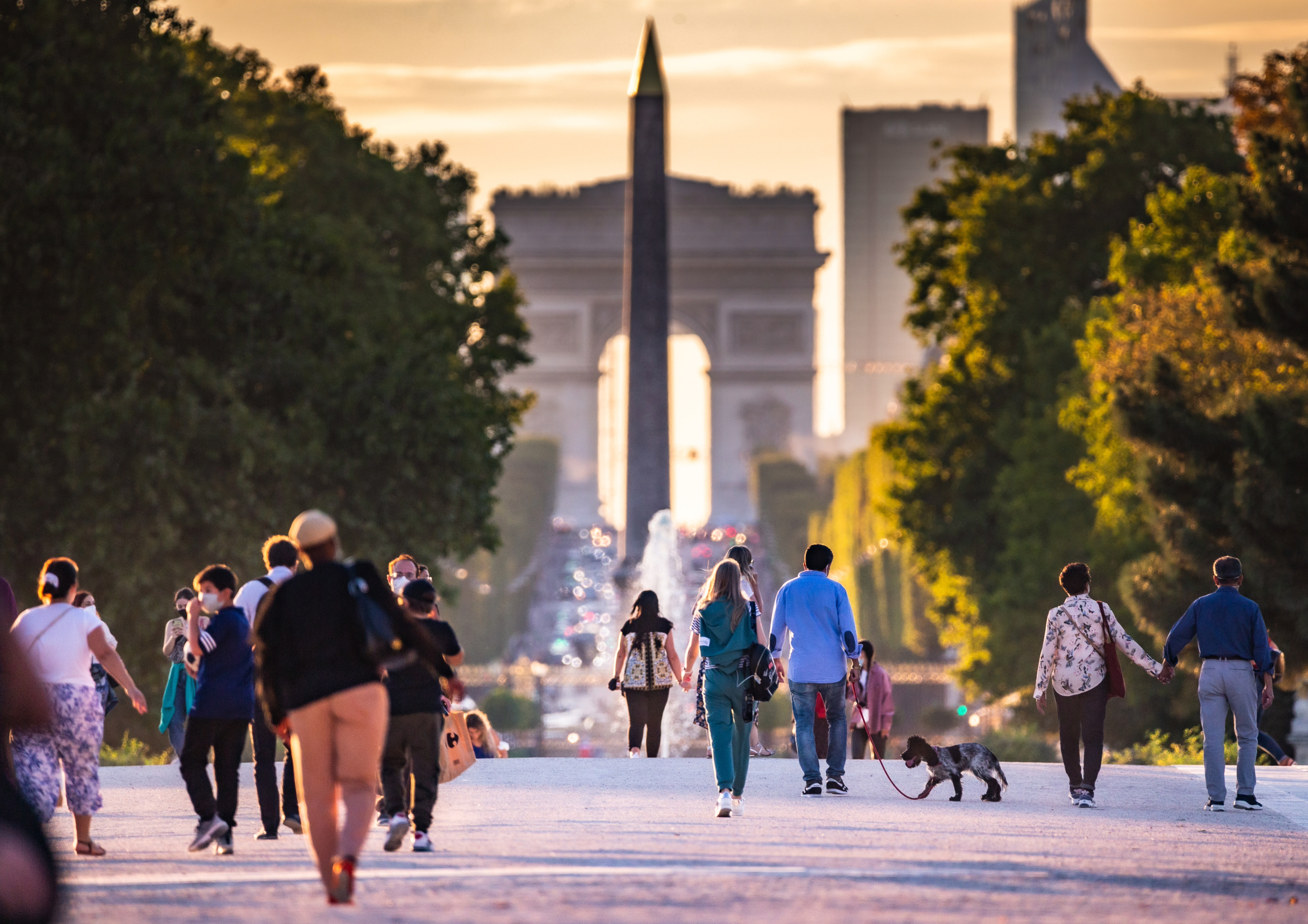People walking and holding hands in front of the Arc de Triomphe during sunset in Paris, with trees lining the avenue and the Eiffel Tower in the background.
