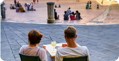 A couple dining outdoors at a table with drinks, overlooking a bustling public square with people sitting and walking around.