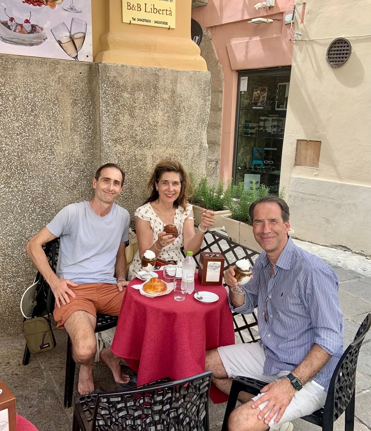 Three adults sitting at an outdoor table enjoying ice cream and desserts. The table has a red tablecloth and is set with glasses, a water bottle, and a napkin holder. The setting appears to be a street cafe with a textured wall and buildings in the background.