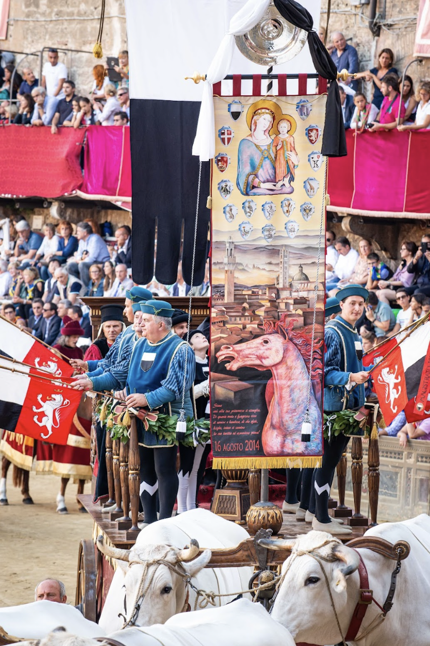 A decorated parade float with a religious banner depicting the Madonna and Child, shield crests, and a mythical horse, surrounded by people in traditional costumes and onlookers at a cultural festival.
