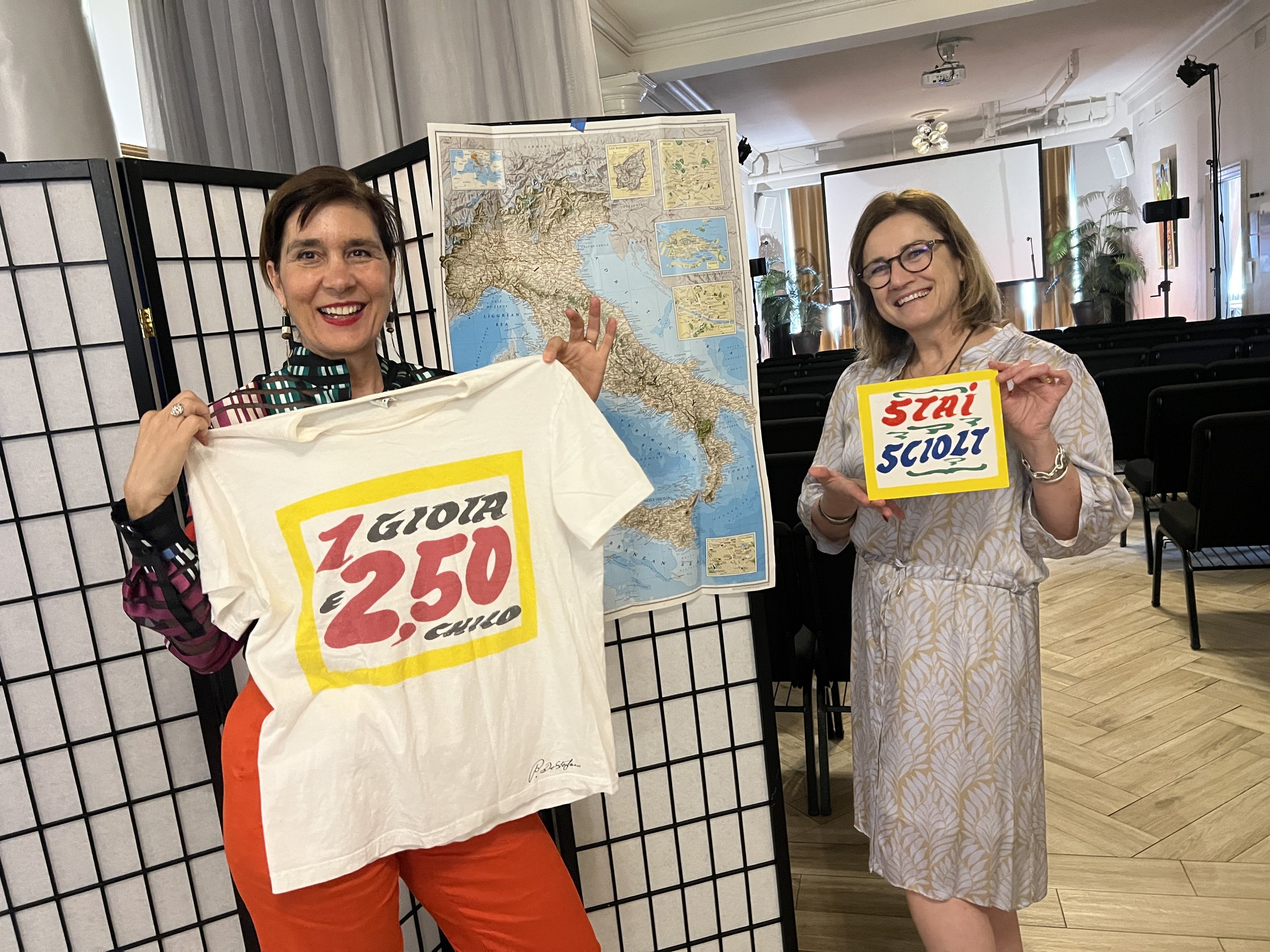 Two women smiling beside a map of Italy at an event.