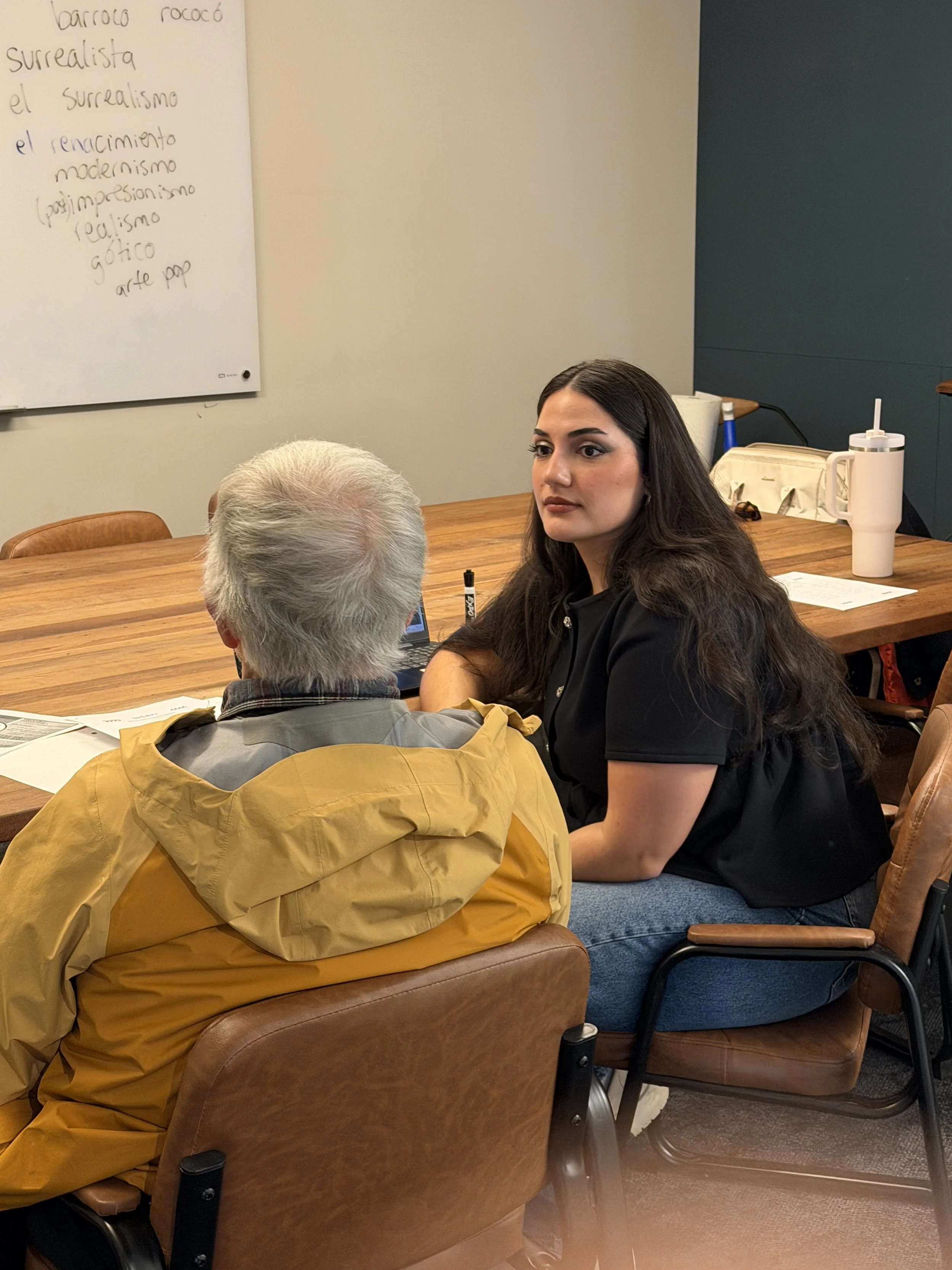 Two people discussing a lesson beside a Spanish-language whiteboard.