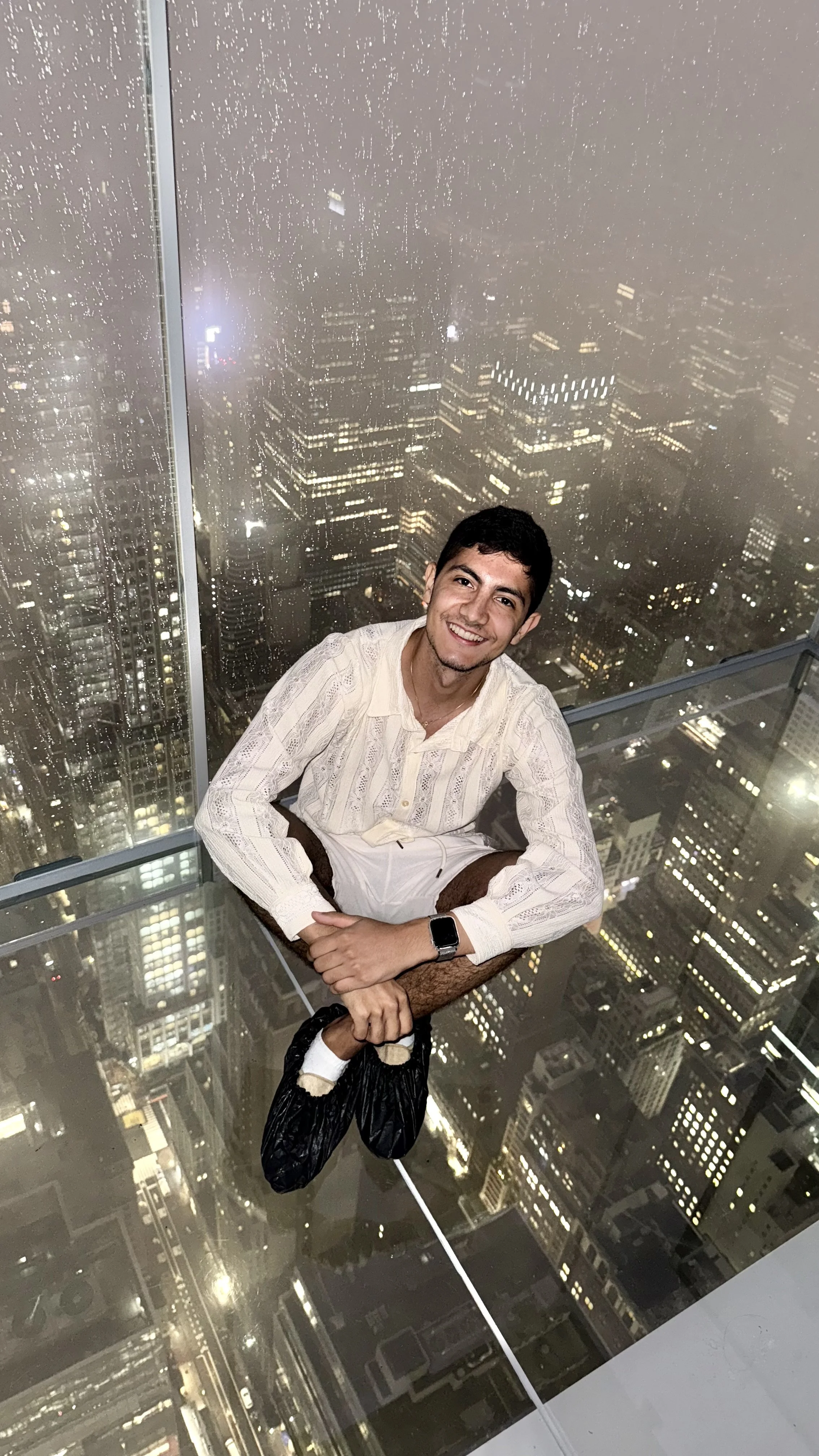 A man sitting on a glass floor above a city at night.