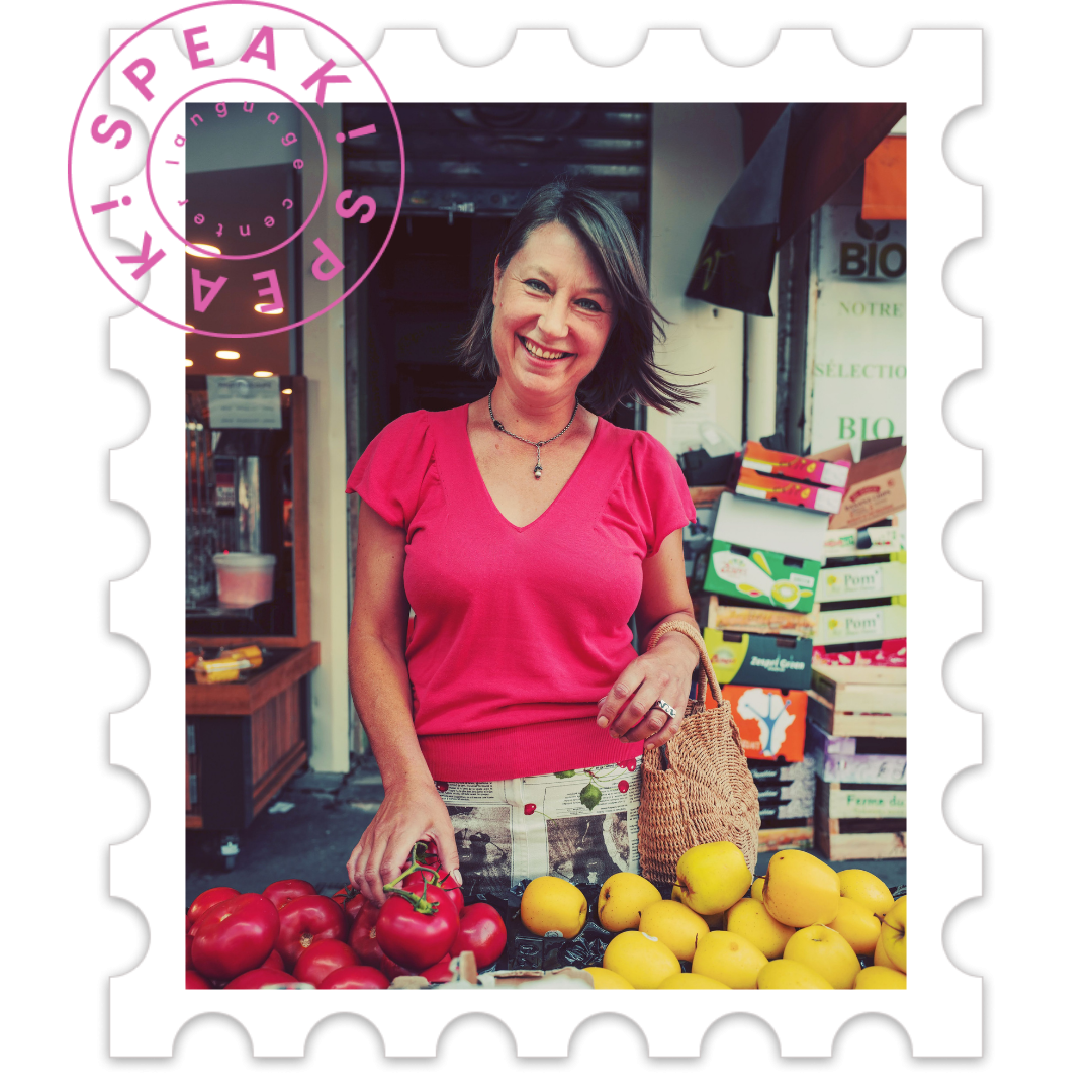 A woman at a farmers market stands behind a produce stall with tomatoes and apples, smiling at the camera.
