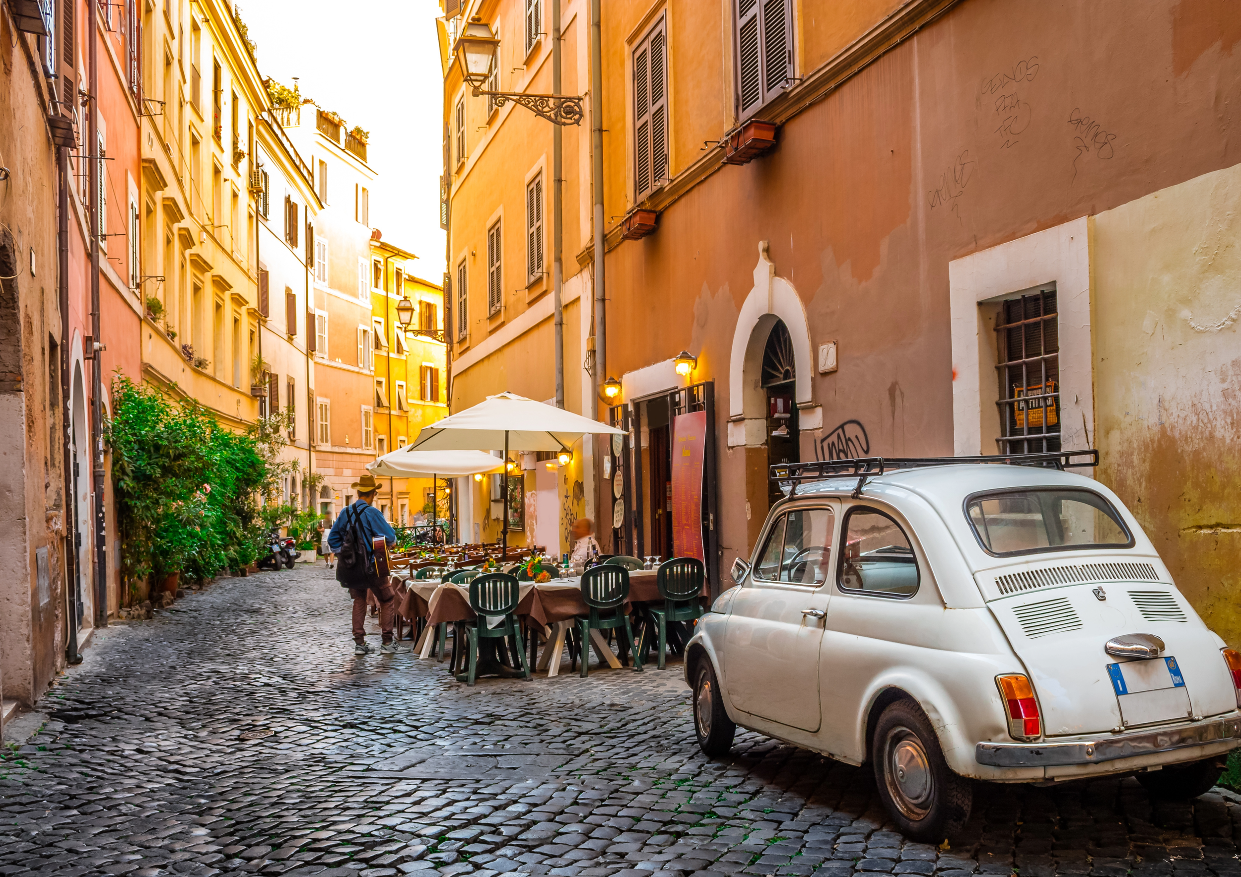 A narrow European cobblestone street with outdoor dining tables covered by umbrellas, colorful buildings, and a vintage white car parked on the side.