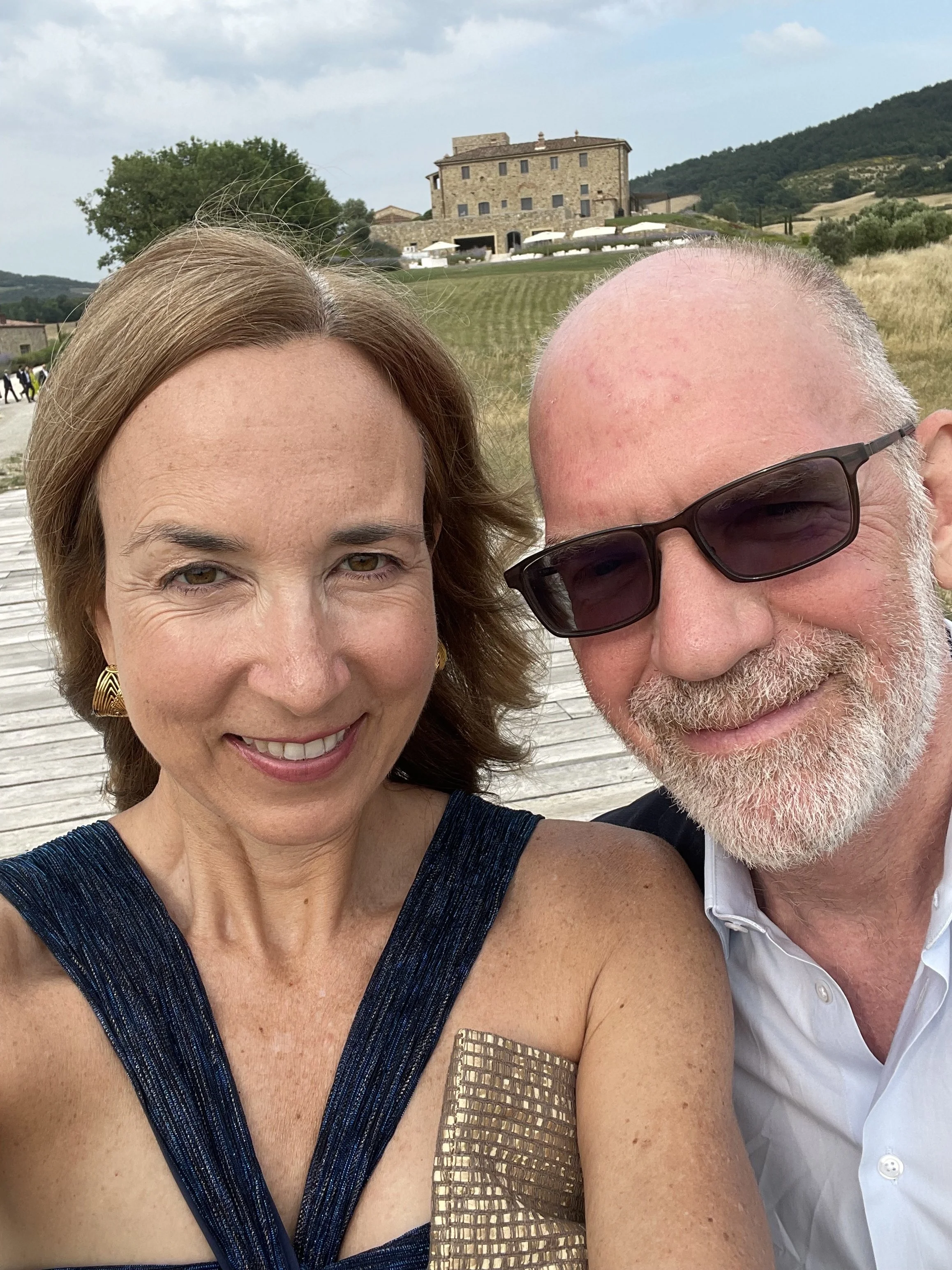 A smiling woman with brown hair wearing a navy dress with gold accents and gold earrings, standing next to a man with sunglasses, a white shirt, and a beard. They are outdoors with a historic stone building, grassy hills, and mountains in the background.