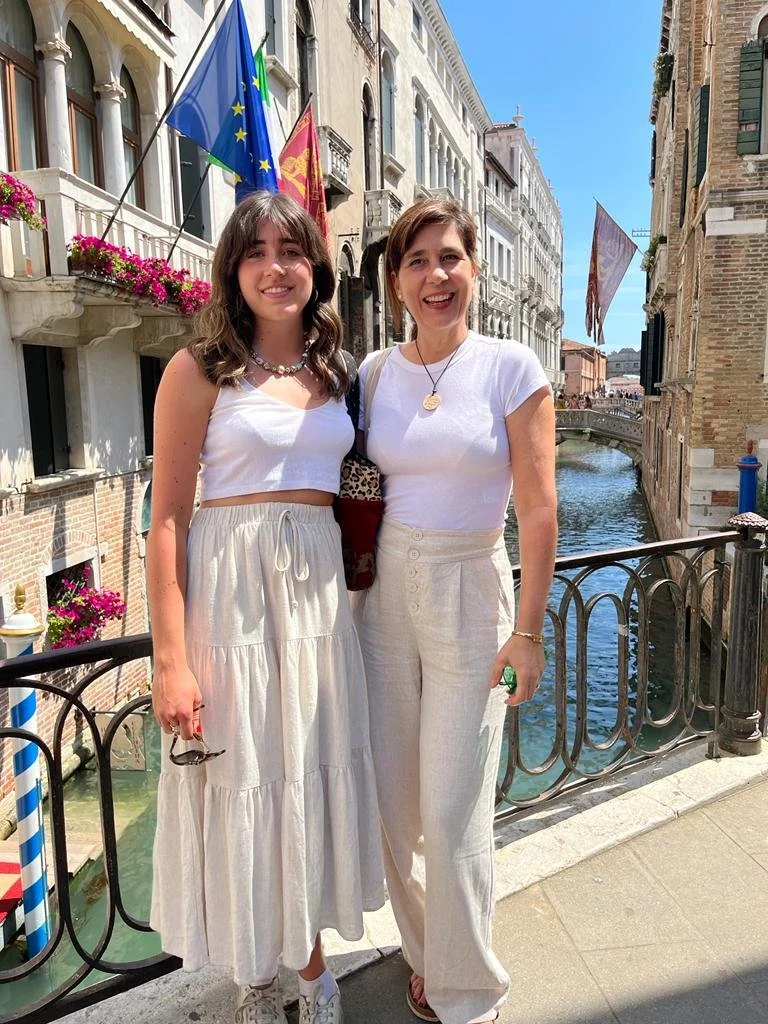 Two women standing on a bridge in Venice, Italy, smiling for the photo, with canals and historic buildings in the background, one holding sunglasses.