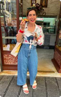 A woman standing outside a shop, smiling and holding a hot dog in one hand and a paper bag in the other. She is wearing a white blouse with colorful patterns, high-waisted blue jeans, and white sandals, with a yellow tote bag over her shoulder.