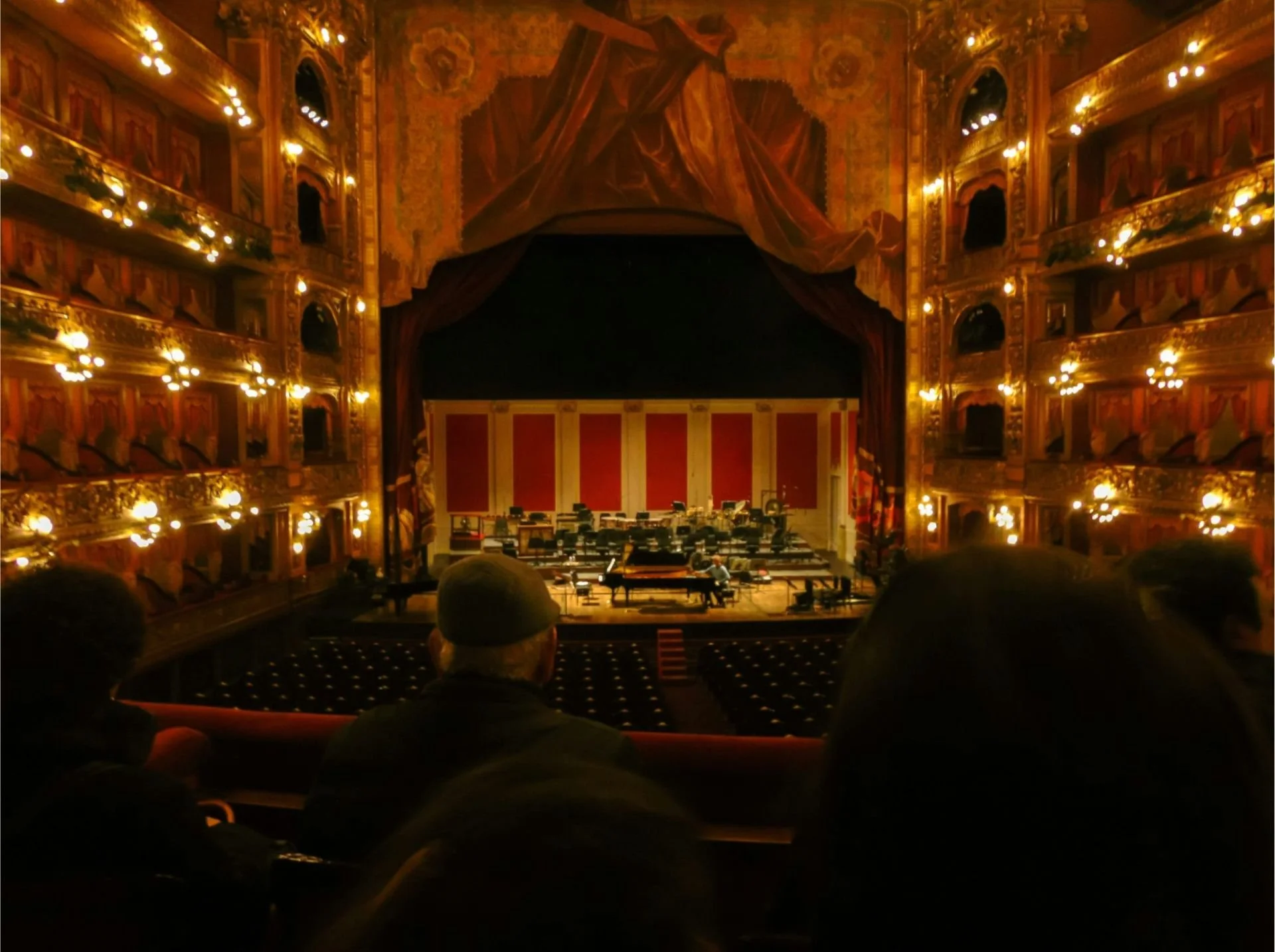 Interior view of a grand theater auditorium with ornate balconies and gold detailing, stage set for a musical performance with chairs, music stands, and a piano, with audience members seated in the foreground.