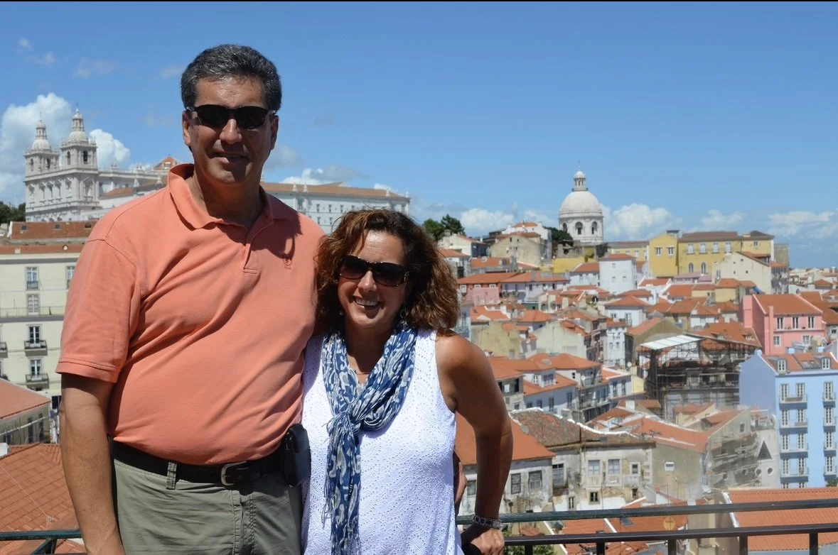 A man and woman with sunglasses smiling on a rooftop overlooking a city with orange roofs and historic buildings under a blue sky.