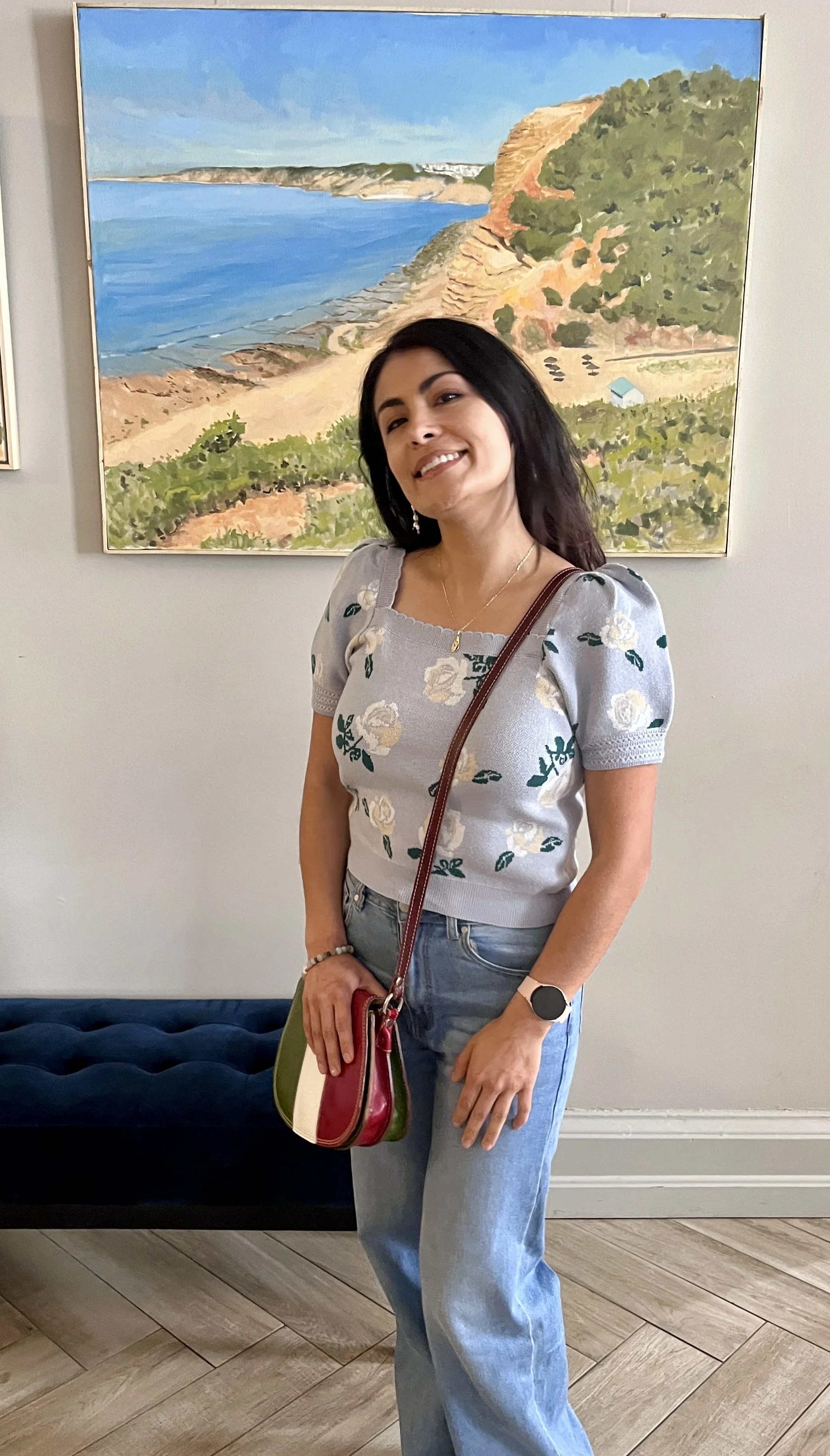 A woman standing indoors in front of a painting of a coastal landscape, smiling and wearing a floral sweater, jeans, and a watch.
