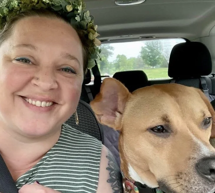A woman with a flower crown smiling next to a tan dog with one ear up inside a car.