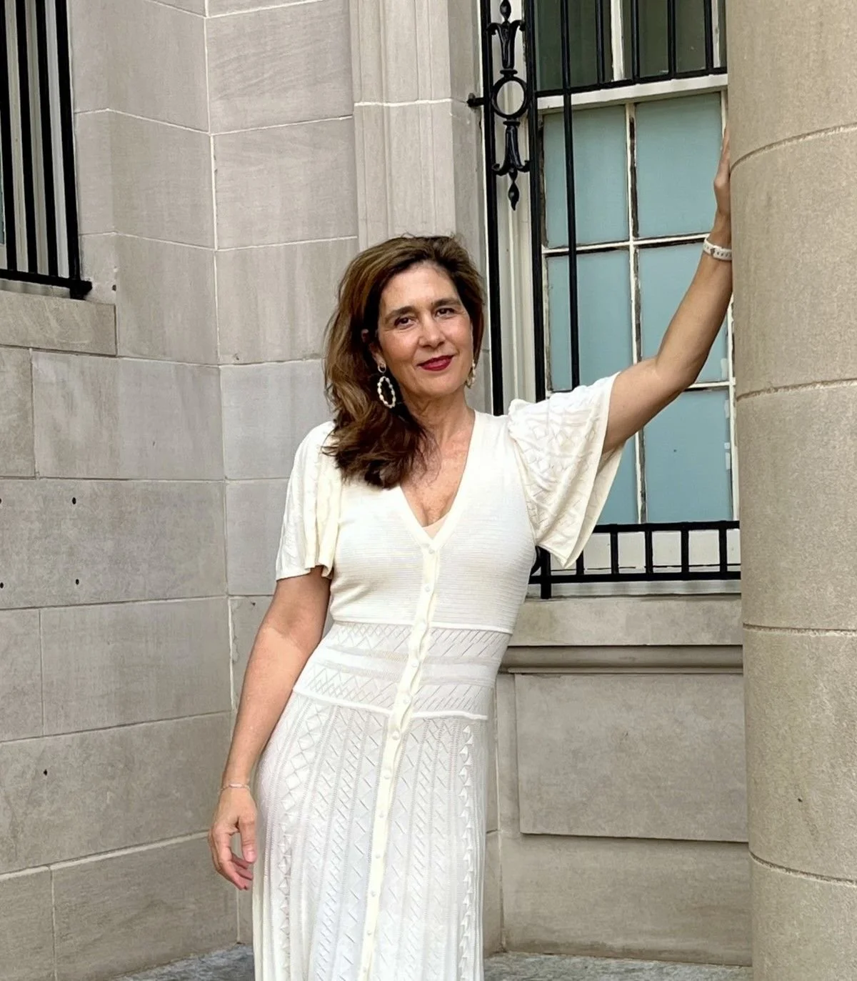 A woman in a white dress standing outside near a stone building with a window and a metal fence.