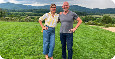 A smiling couple standing on a grassy field with mountains and trees in the background during daytime.