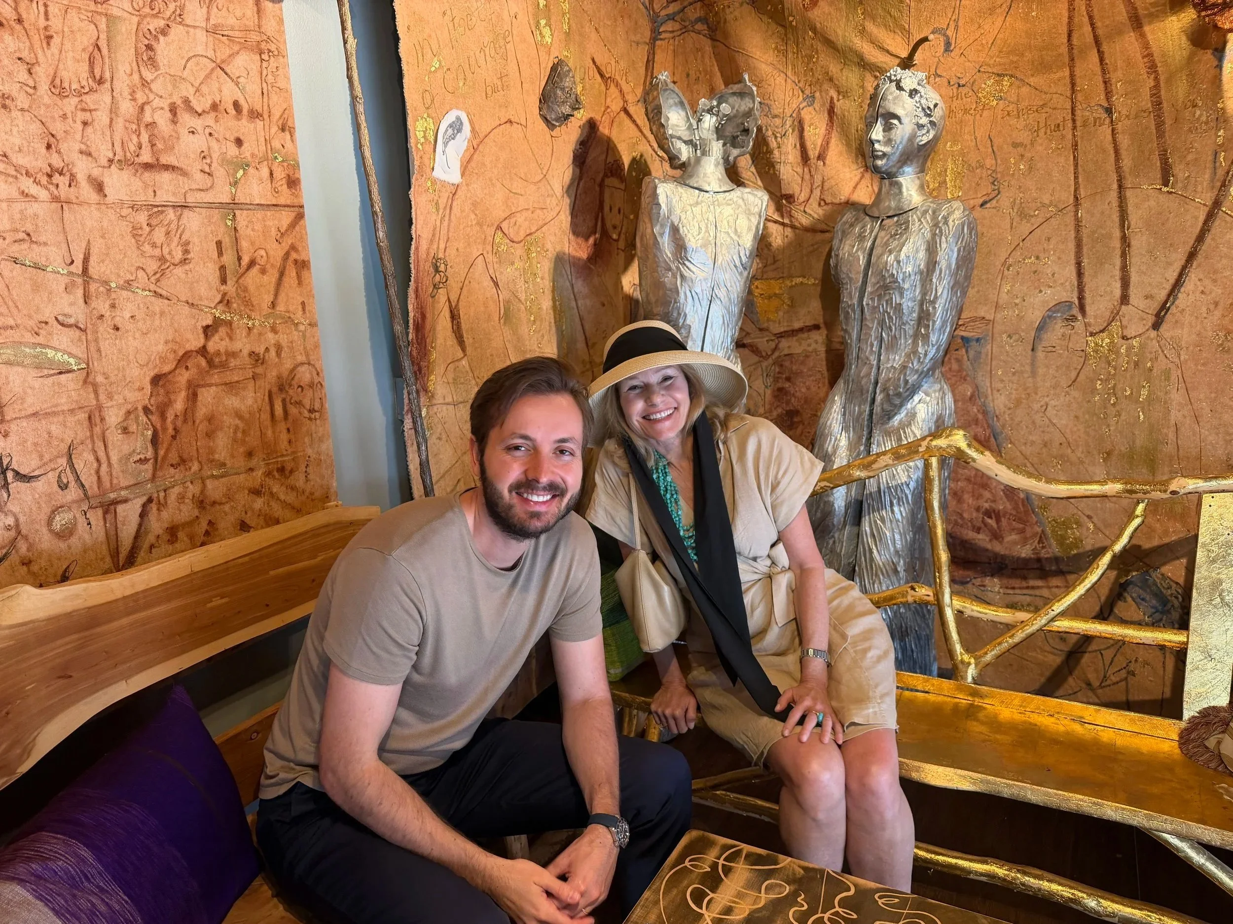 Two people smiling on a bench in front of artwork.
