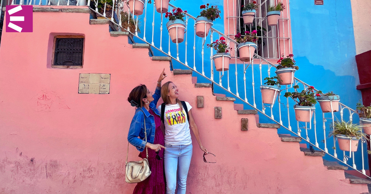 Two women standing under a staircase decorated with hanging pink flower pots on a pink and blue wall.