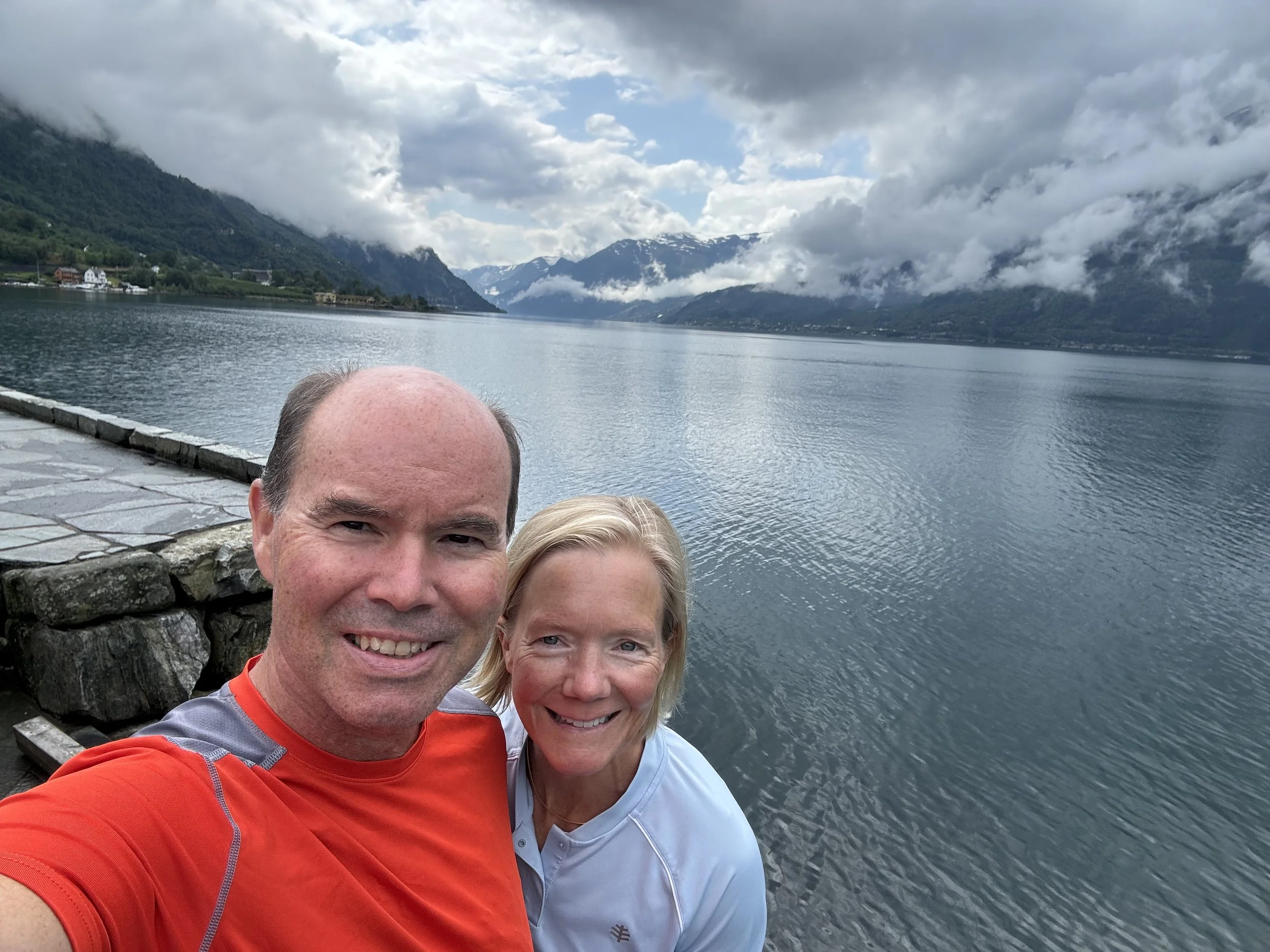 A smiling man and woman taking a selfie by a lake with mountains and clouds in the background.