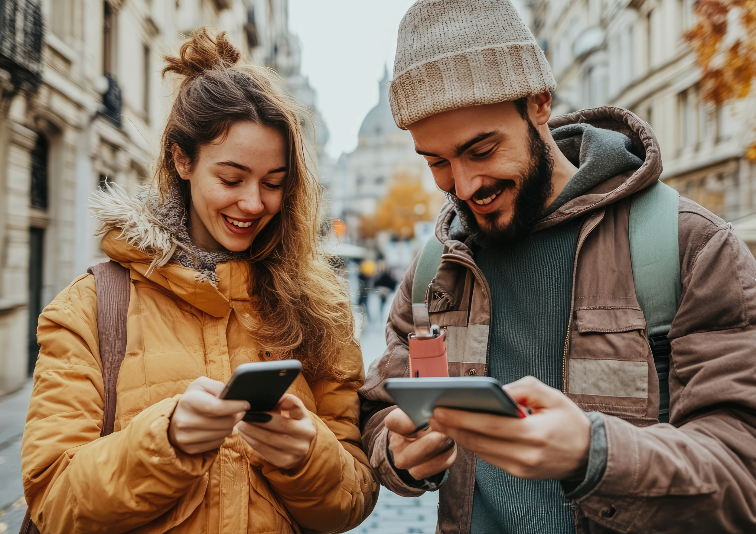 A young woman and man in autumn clothing are smiling and looking at their smartphones while standing on a city street.