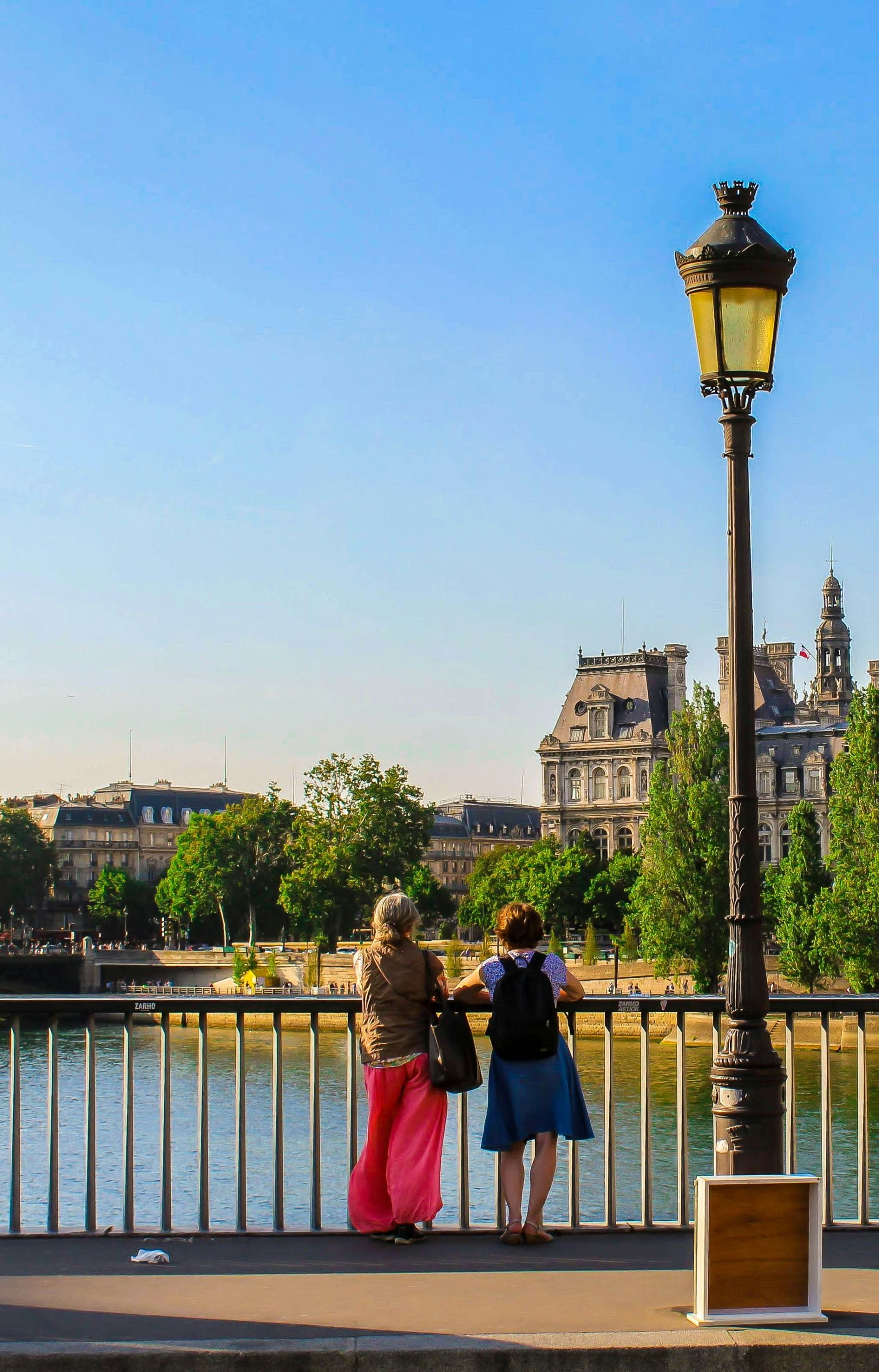 Two women standing on a riverside promenade in Paris, France, looking at the river with historic buildings in the background and a vintage lamp post in the foreground.