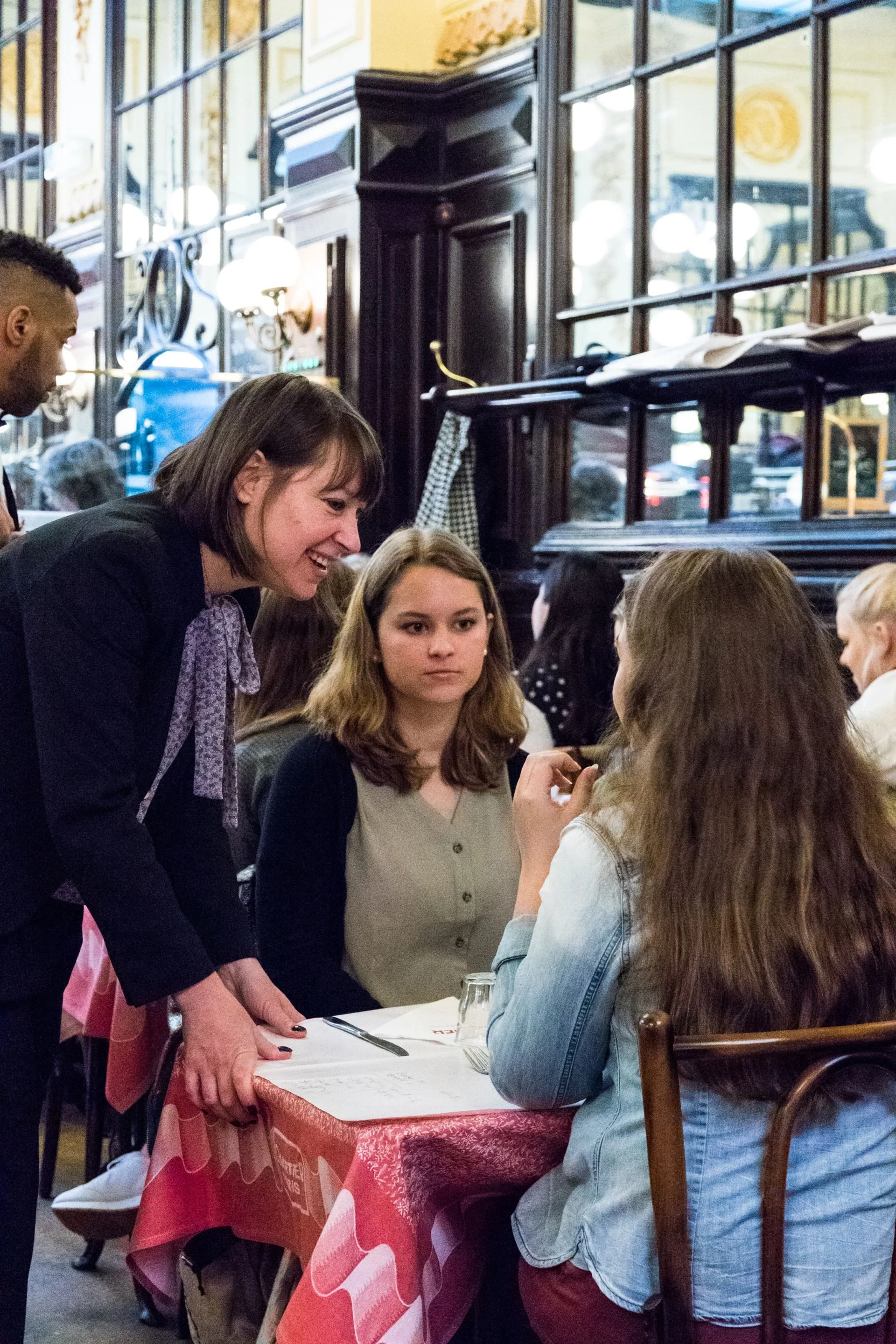 A waitress takes an order from two women seated at a restaurant table, with other patrons visible in the background.