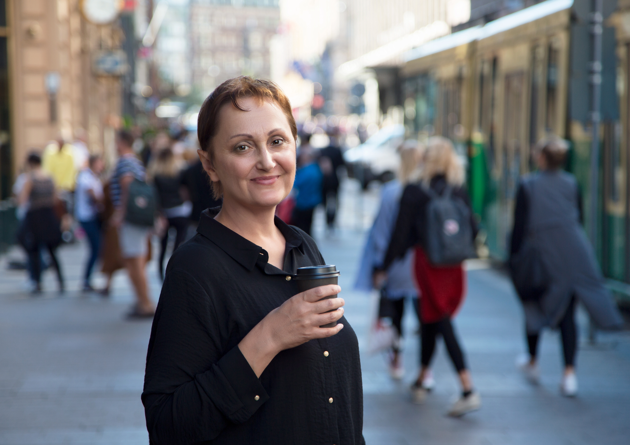 A woman with short brown hair holding a coffee cup on a busy city street with pedestrians and a green trolley in the background.