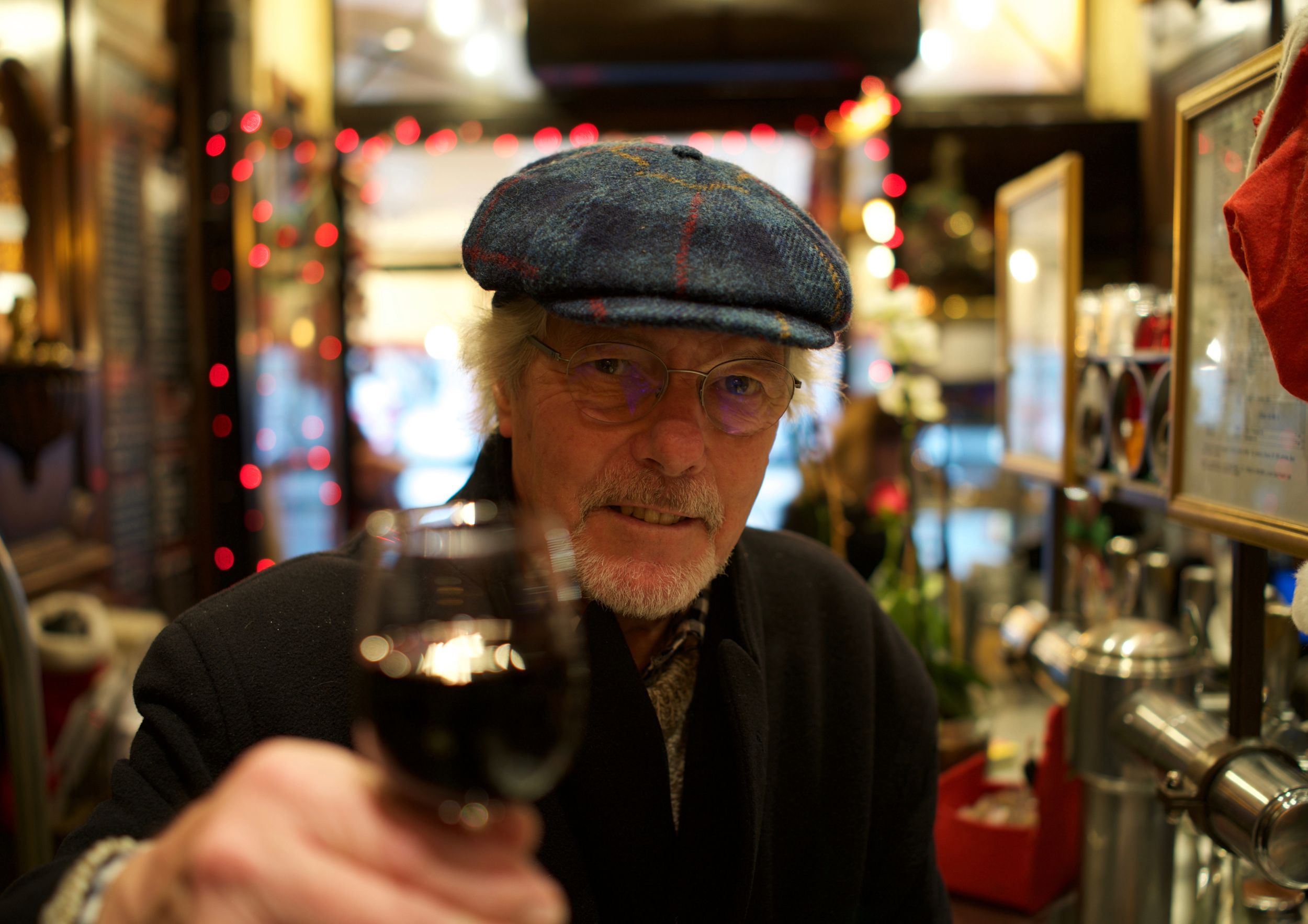 An elderly man with blonde hair, glasses, and a beard, wearing a blue plaid flat cap, holding a glass of red wine, in a cozy, decorated indoor space.