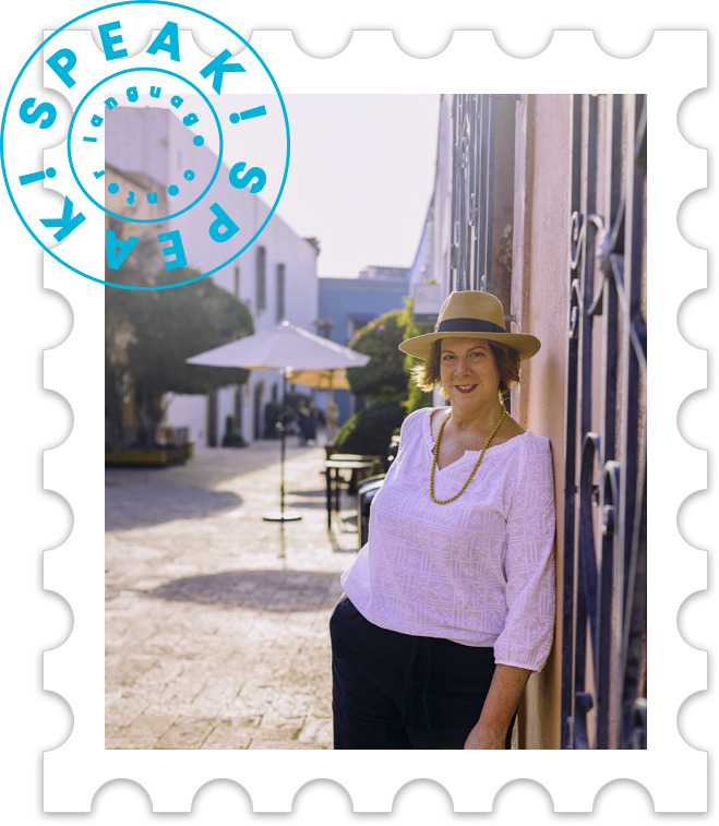Woman in white blouse and hat leaning against a wall on a sunny outdoor street with umbrellas and buildings in the background.