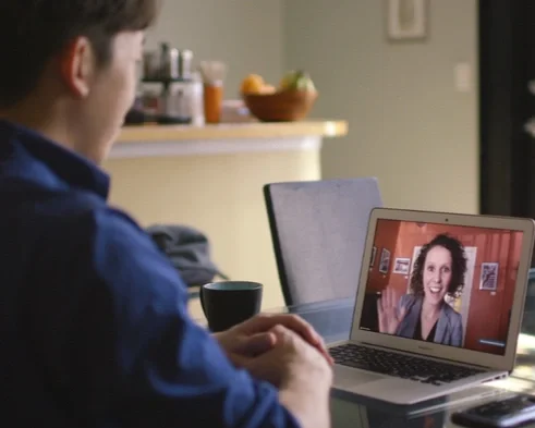 A man in a blue shirt participating in a video call on a laptop, with a woman smiling and waving on the screen.