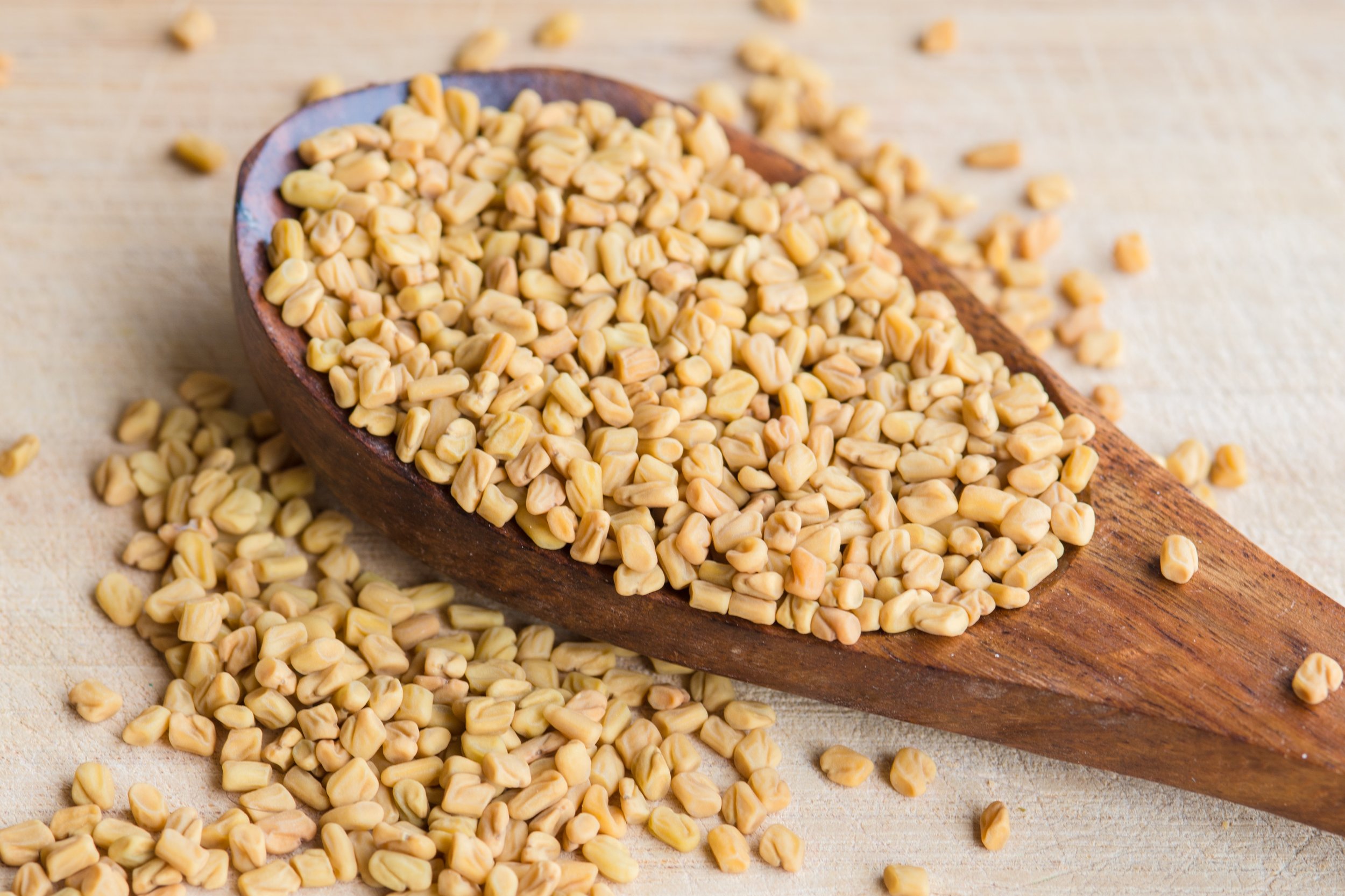 Wooden spoon filled with dried fenugreek seeds, some spilling onto a light-colored surface.