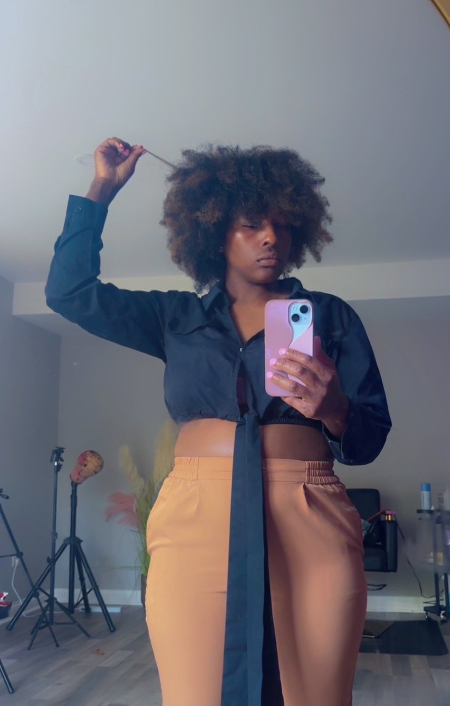 Woman with curly hair in a mirror selfie, holding a pink phone case, wearing a black crop top with long sleeves and high-waisted beige pants, in a room with studio equipment in the background.