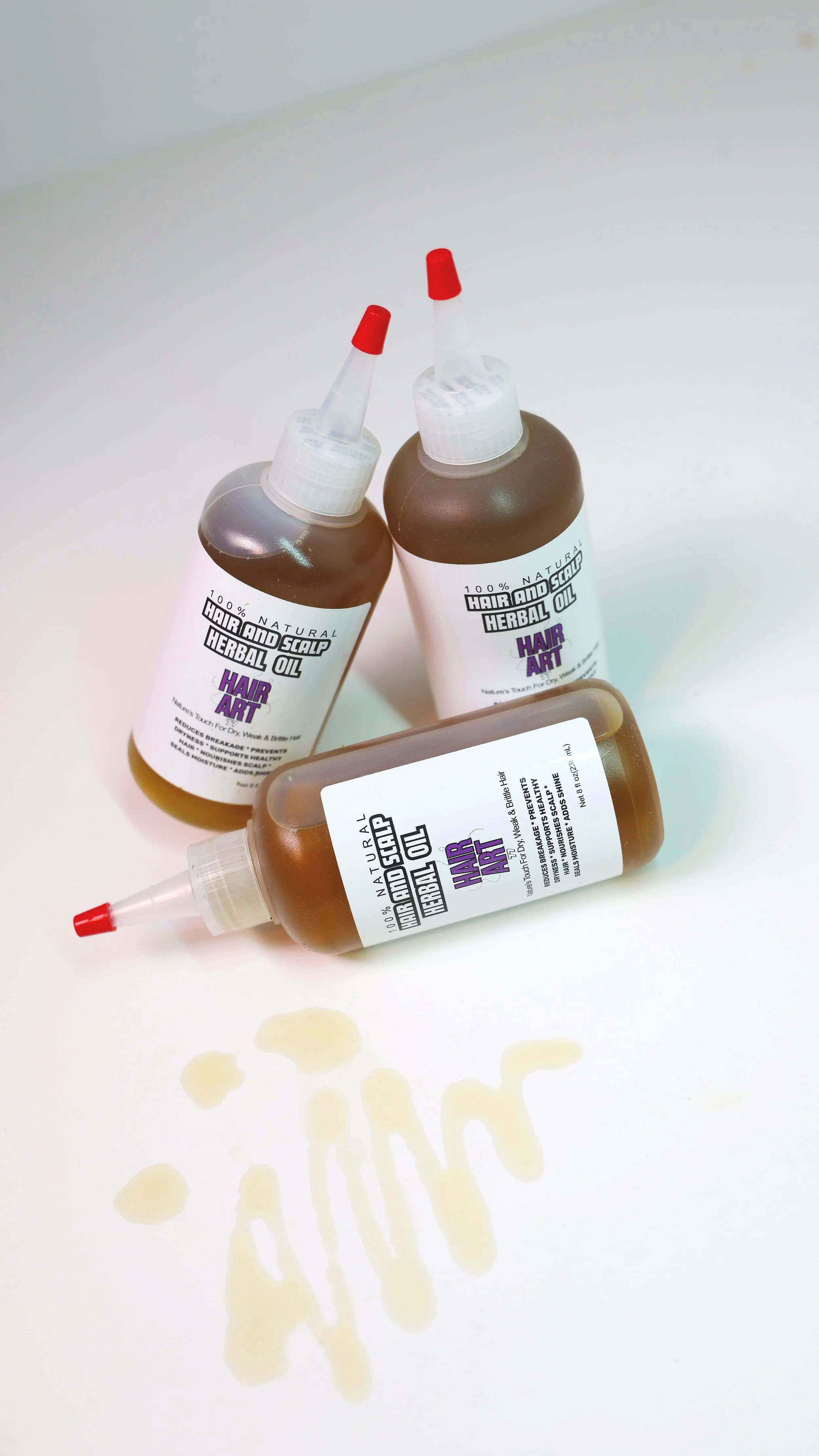 Three bottles of herbal scalp and hair oil with a spilled amount on a white surface.