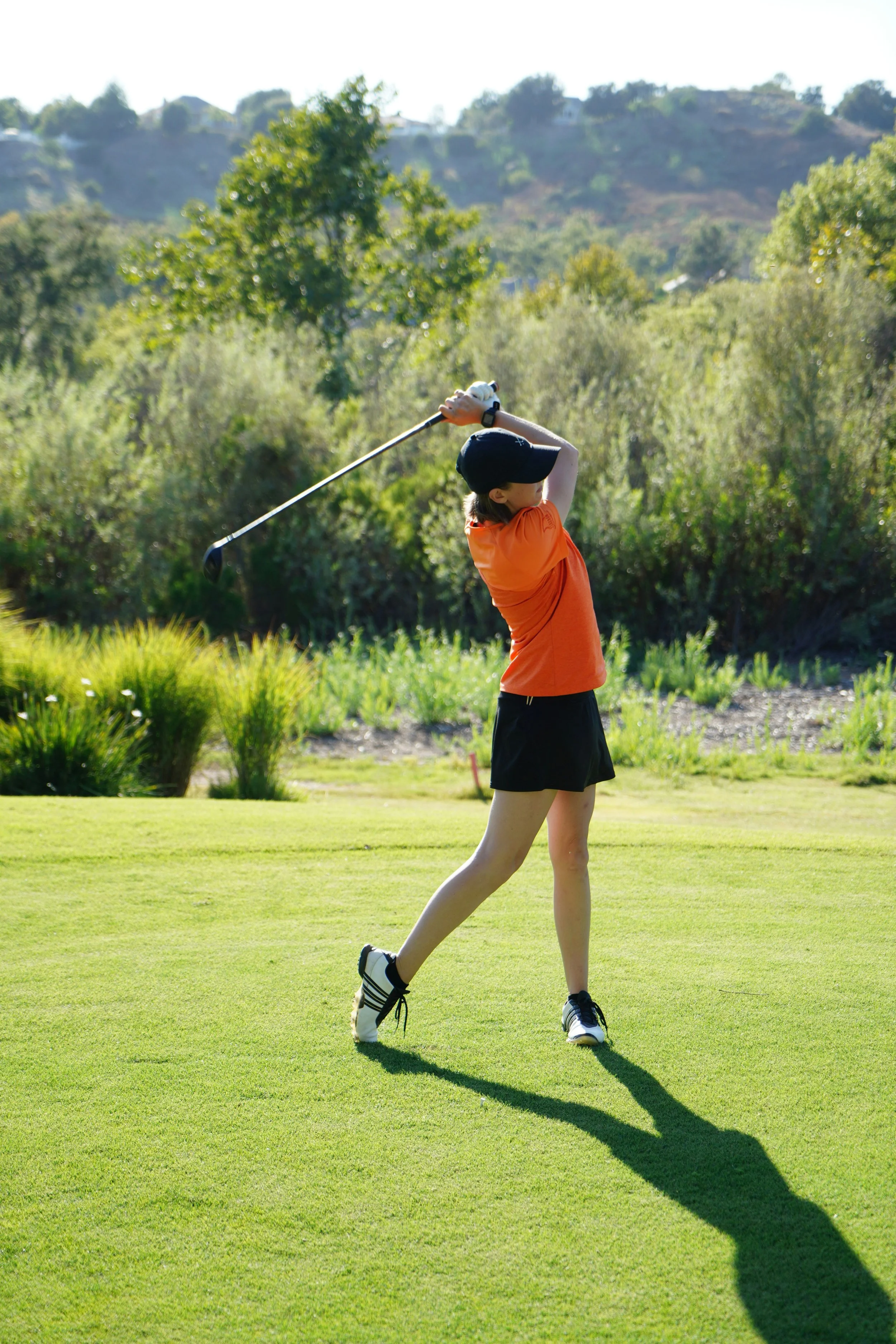 A female golfer in a red shirt, black shorts, and a black cap, swinging a golf club on a golf course.