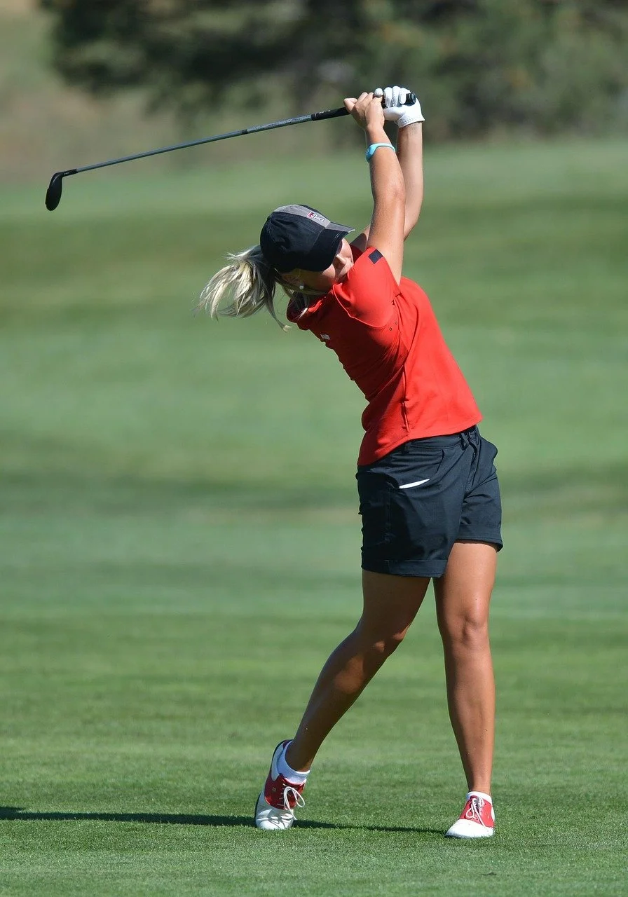 A female golfer in a red shirt and black shorts swinging a golf club on a golf course.