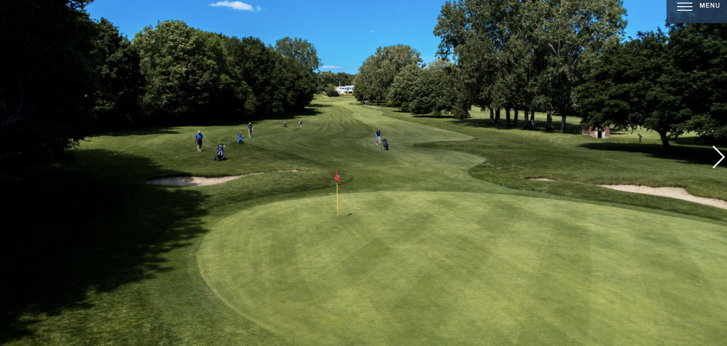 A view of a golf course with a putting green in the foreground, a flagstick with a red flag, and players in the distance near trees on a sunny day.