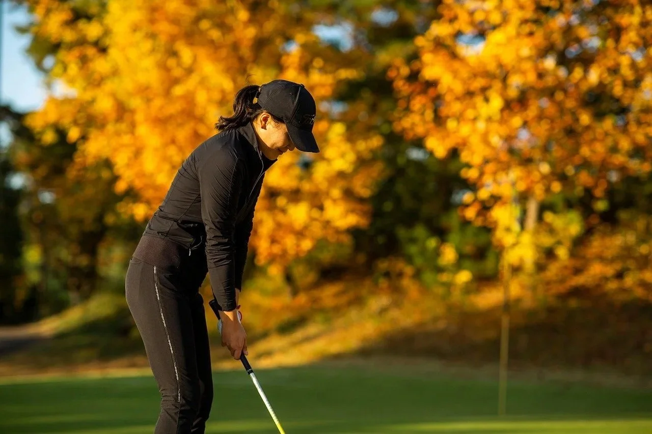 A woman dressed in black, playing golf on a course with autumn-colored trees in the background.