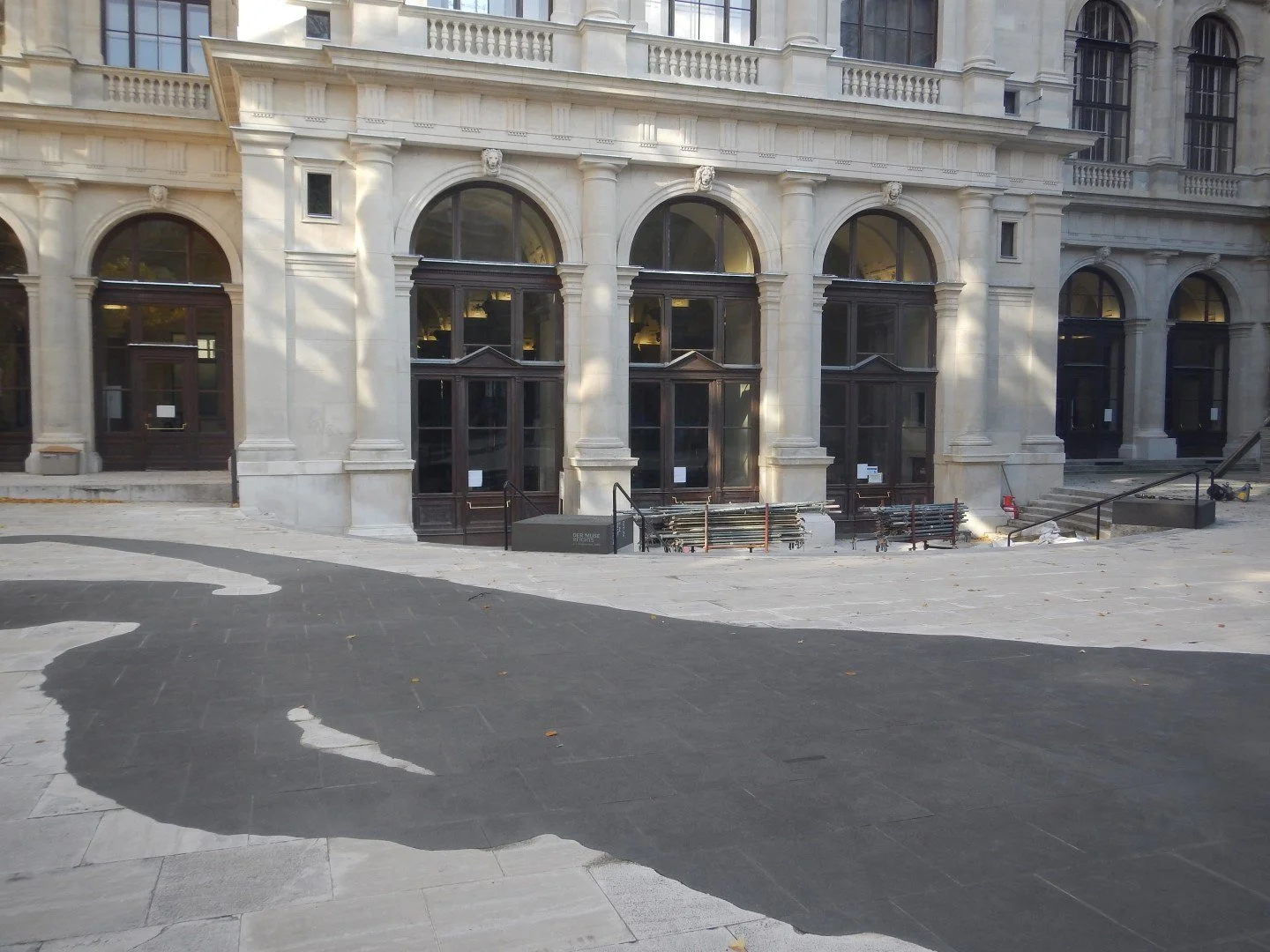 Ein leerer, gepflasterter Platz vor einem historischen Gebäude mit großen Fenstern und klassischen Säulen, vermutlich in einer Stadt.