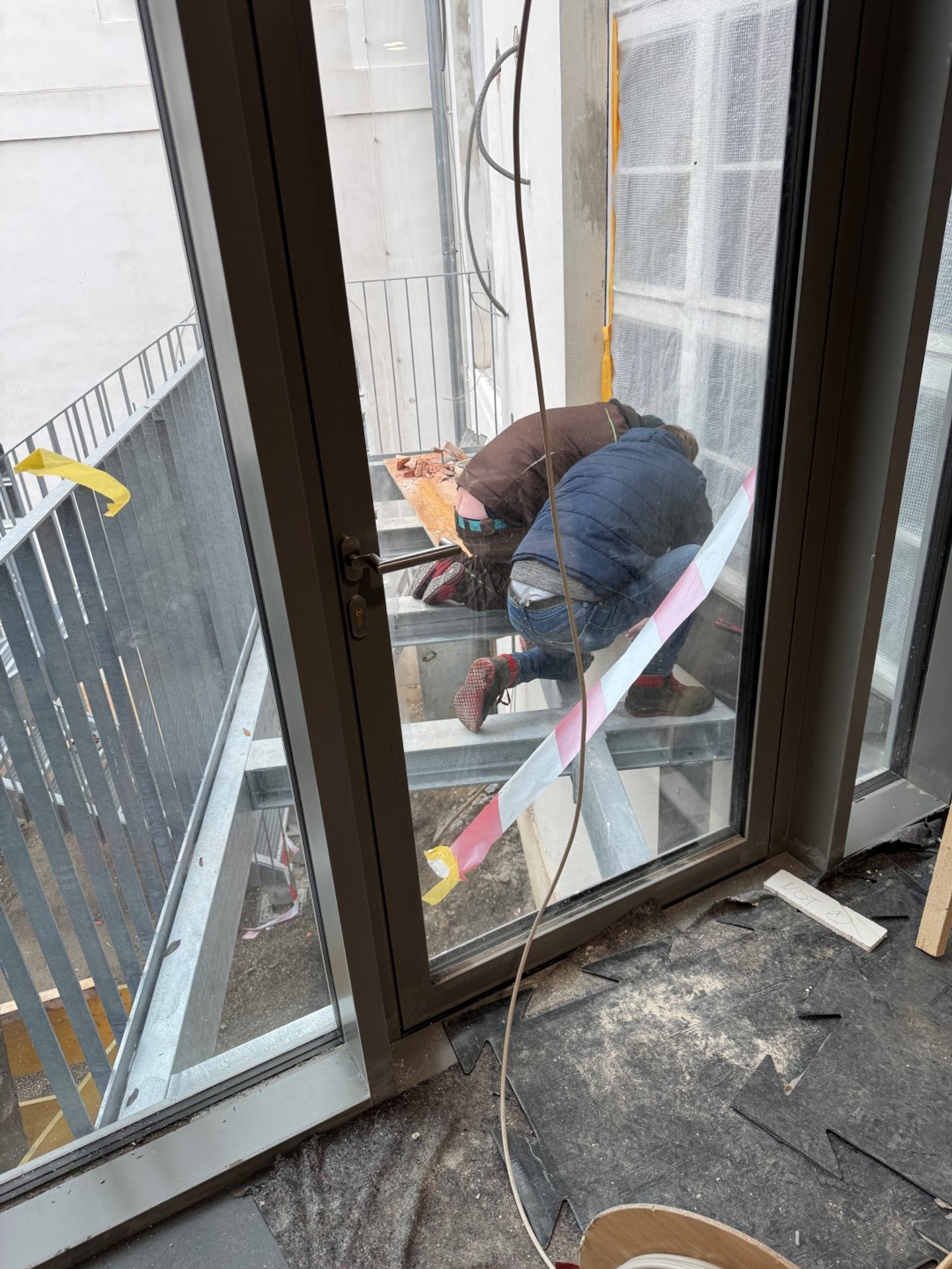 Zwei Bauarbeiter arbeiten auf einer kleinen Baustelle vor einem Glaseingang, mit Schutzband und Baumaterial. Das Bild wird durch eine Fensterscheibe aufgenommen.