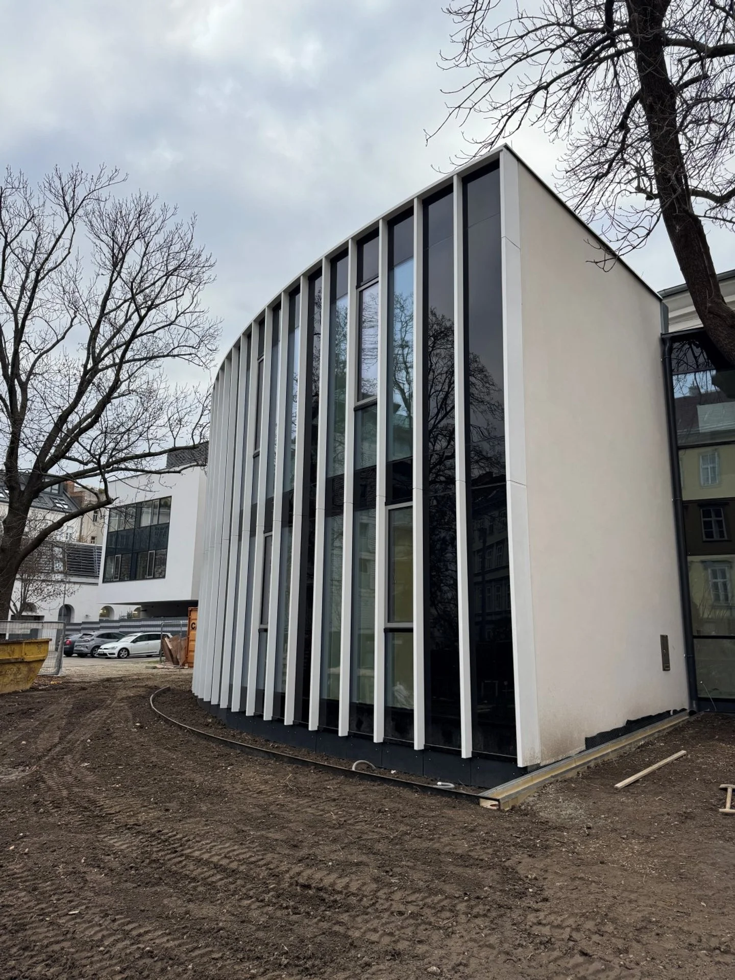 Neues modernes Gebäude unter Bau, mit großen Fenstern und vertikalen weißen Elementen, umgeben von unbefestigtem Boden und Bäumen.