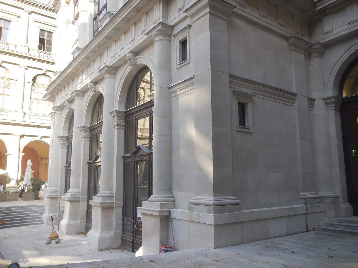 Ein Gebäudeteil mit großen Fenstern, weißen Steinsäulen und Barockelementen an einer klassischen Architektur. Im Hintergrund sind weitere Gebäude zu sehen.