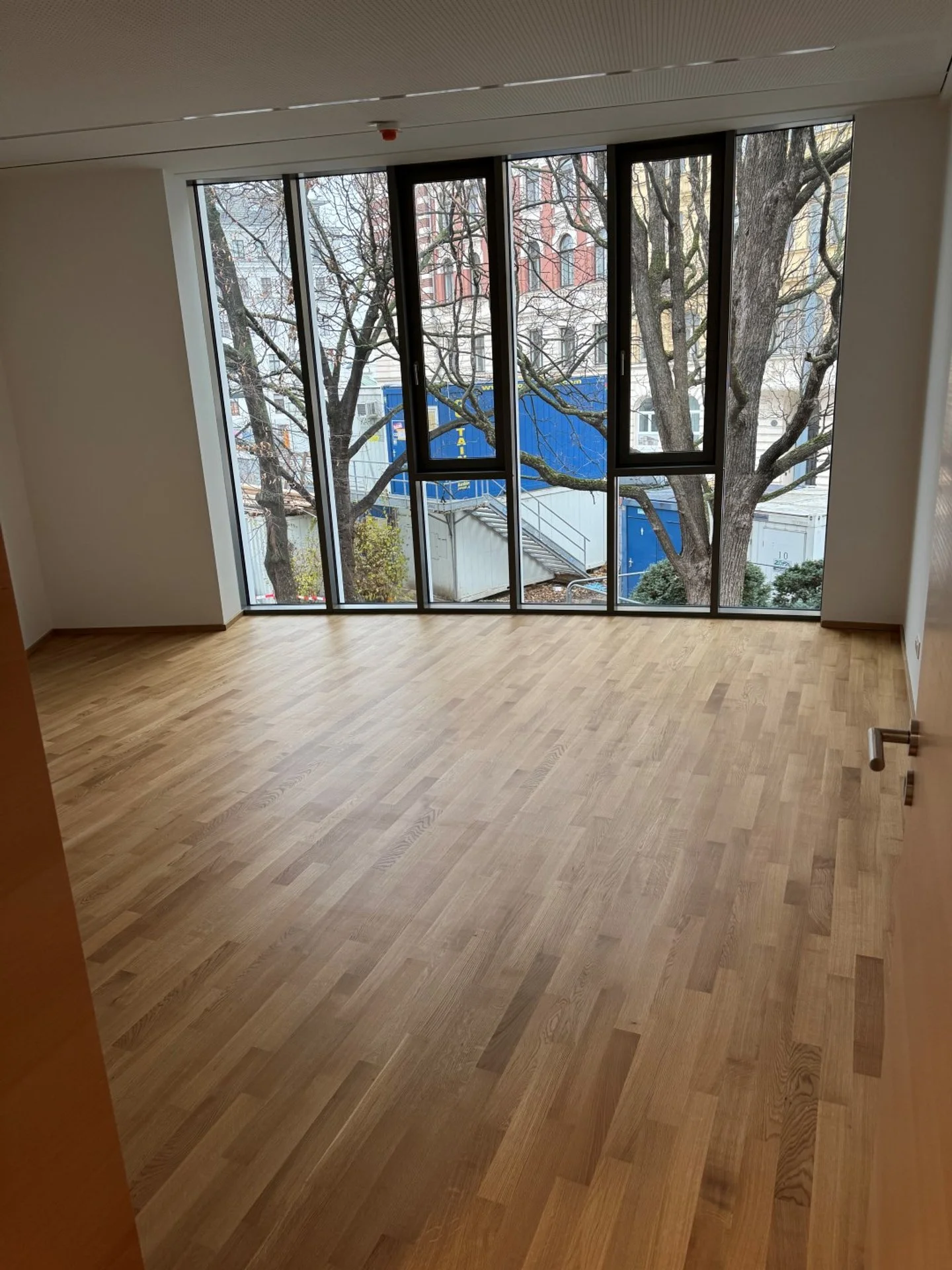 Leeres Zimmer mit großen Fenstern, Holzfußboden und Blick auf Bäume im Freien. Von bmk-projekt umgesetzt in Wien.