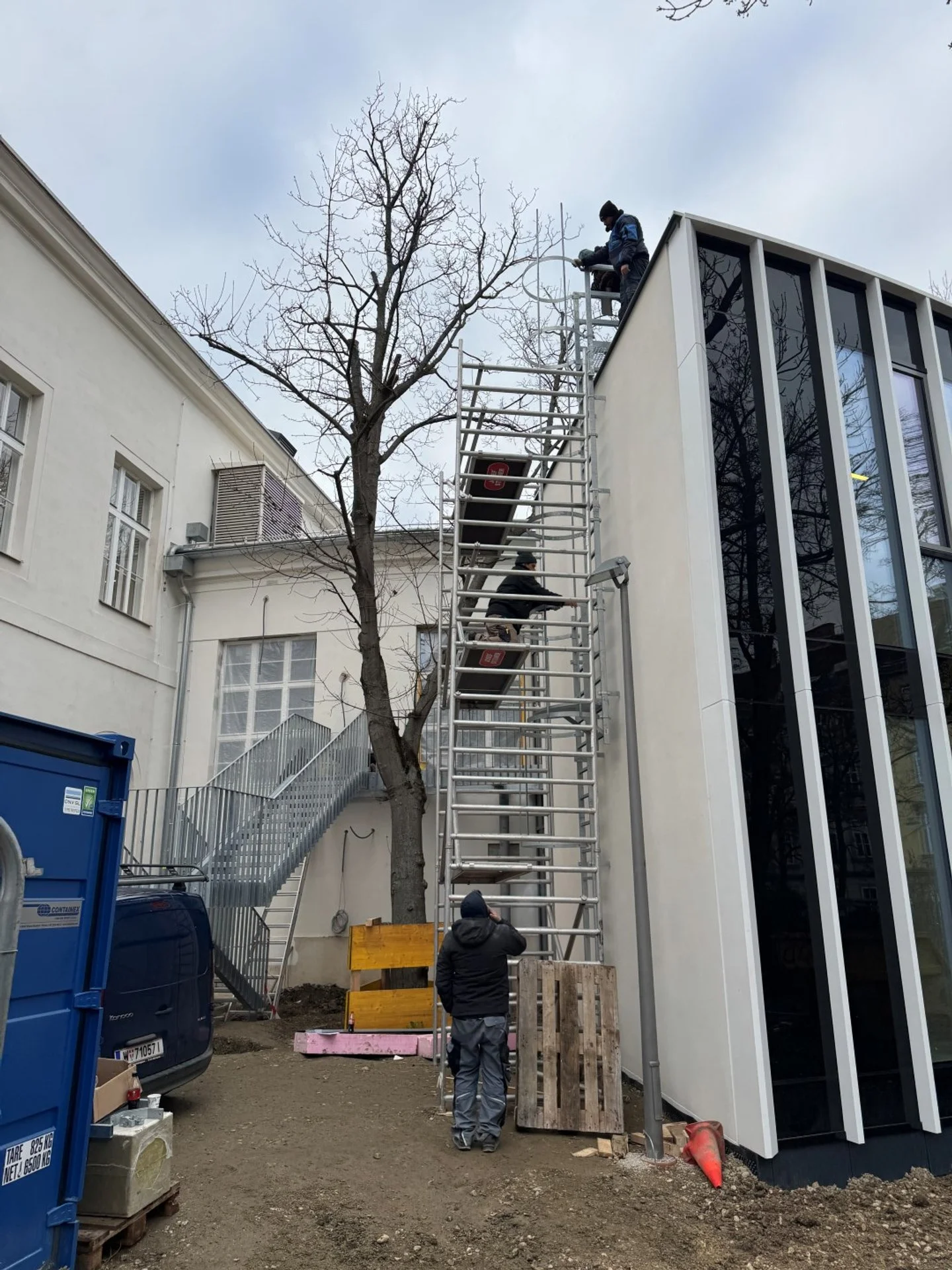 Drei Arbeiter auf einer Baugerüsttreppe vor einem modernen Gebäude, arbeiten an der Dachinstallation bei bewölktem Himmel.