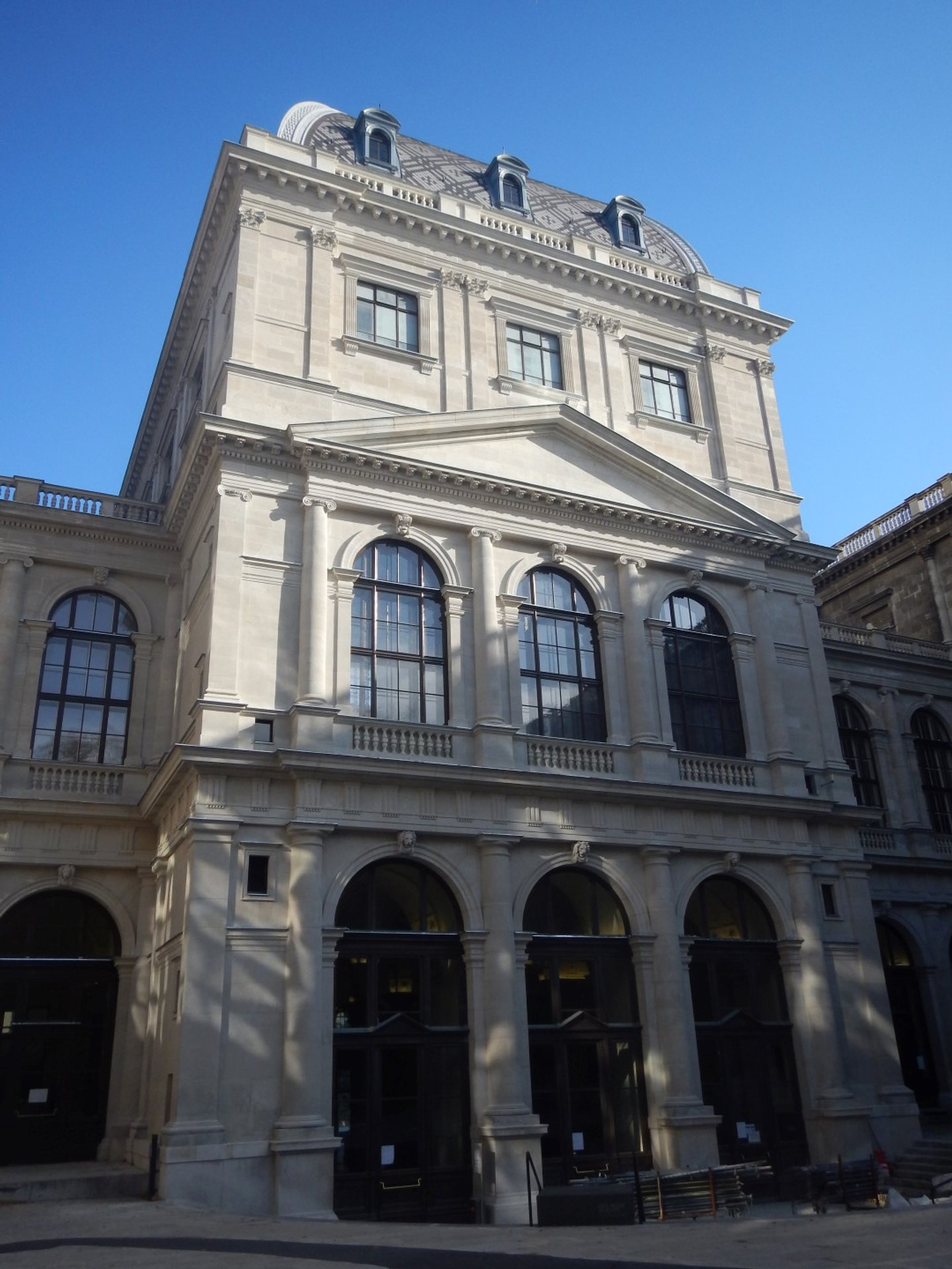 Historisches Gebäude mit klassischer Architektur, große Fenster und detaillierte Fassadenverzierungen, blauer Himmel im Hintergrund.