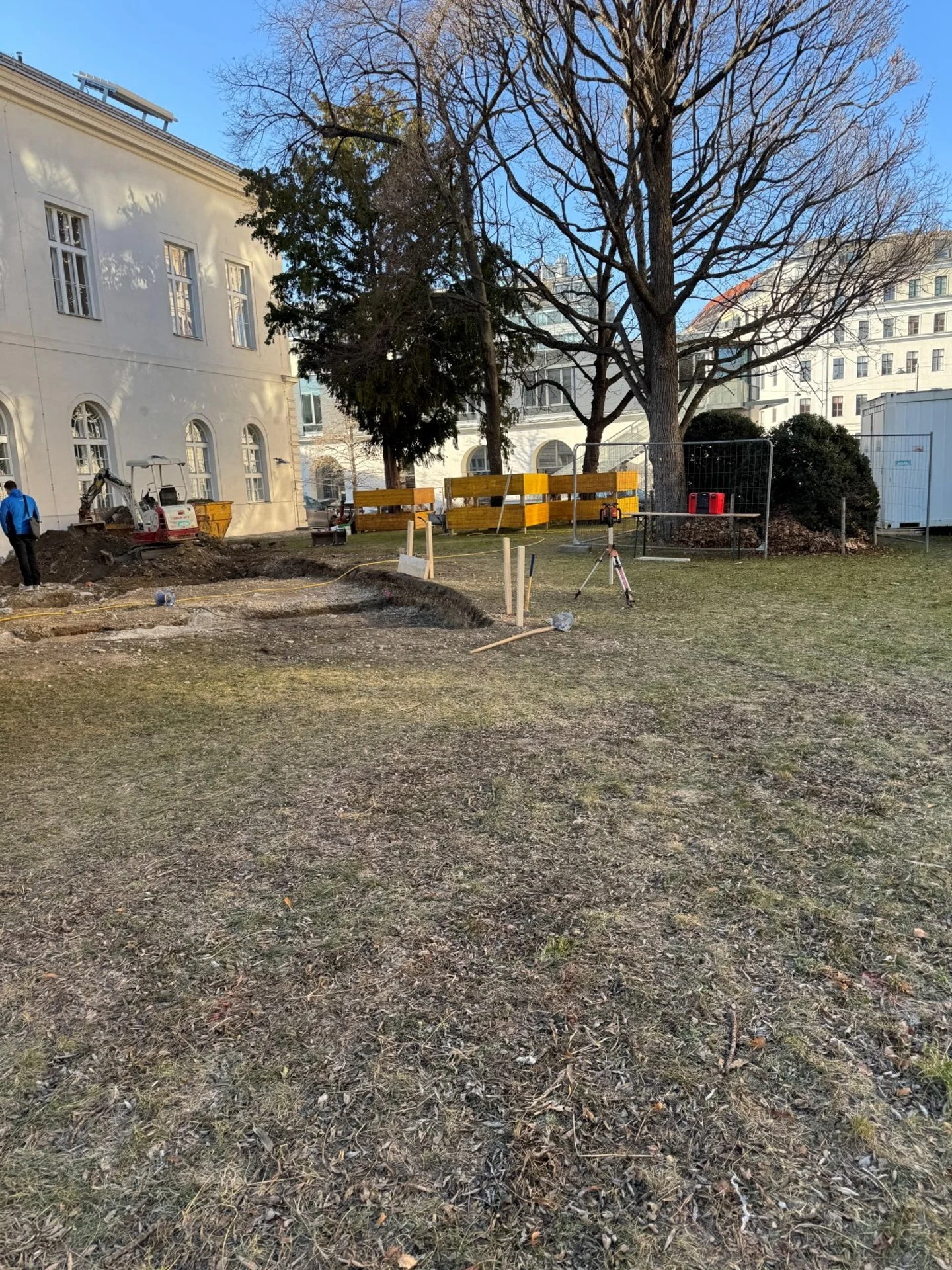 Bauarbeiten in einem öffentlichen Park, aufgerissener Boden, Baugeräte und Bauzäune zu sehen, Bäume und Gebäude im Hintergrund, es ist tagsüber bei schönem Wetter.