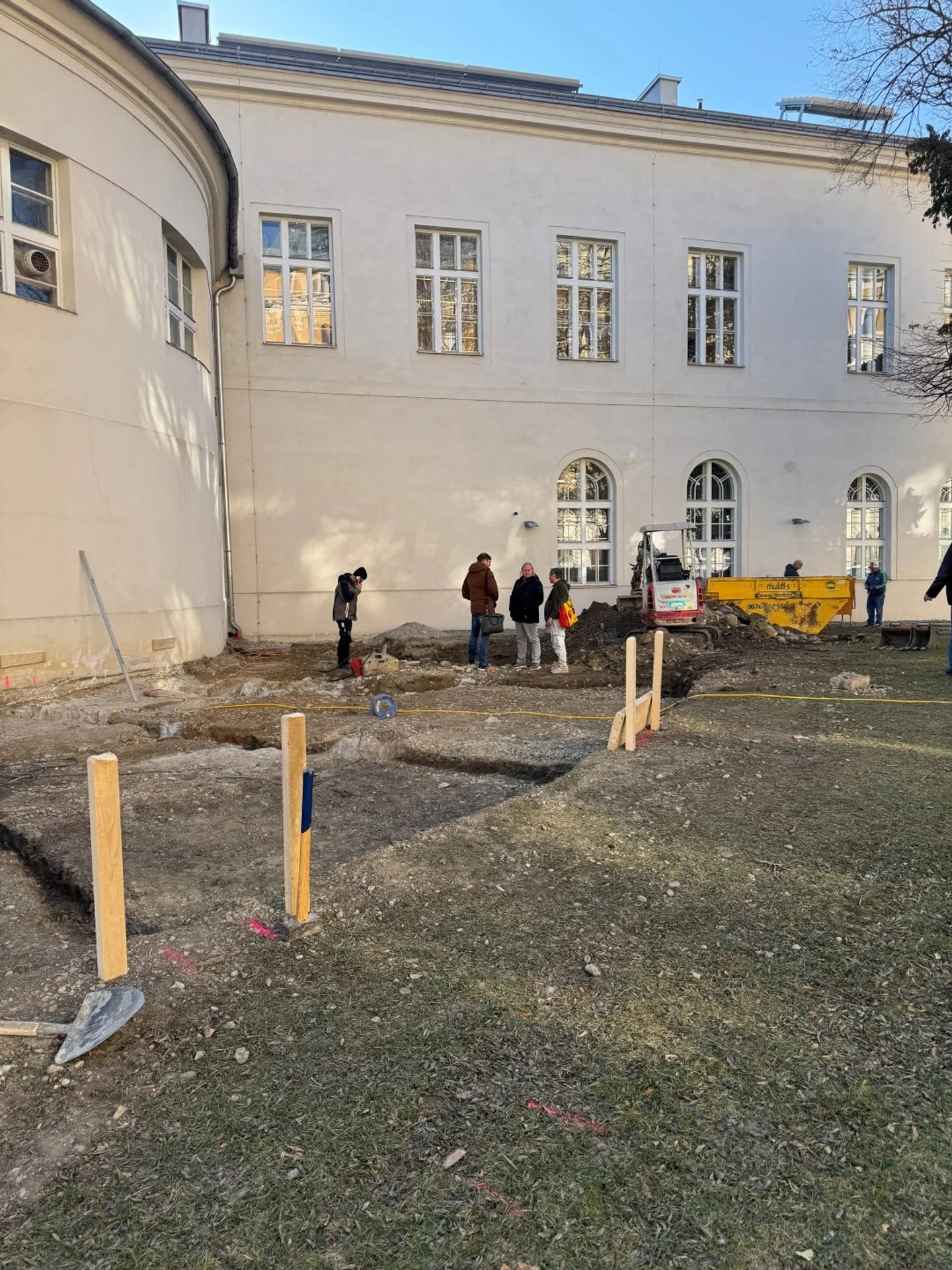 Menschen arbeiten auf einer Baustelle im Freien vor einem weißen Gebäude mit großen, arched und rechteckigen Fenstern. Es gibt Baugeräte, Schaufeln, und Holzpfosten in der Erde.