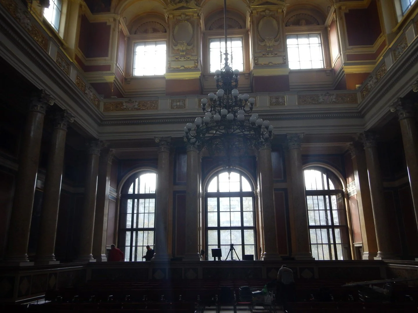 Ein prunkvoller Saal mit hohen Fenstern, großen Säulen, einer großen Kristallleuchter und aufwändiger Deckenverzierung, im Hintergrund drei Menschen und ein Stativ.
