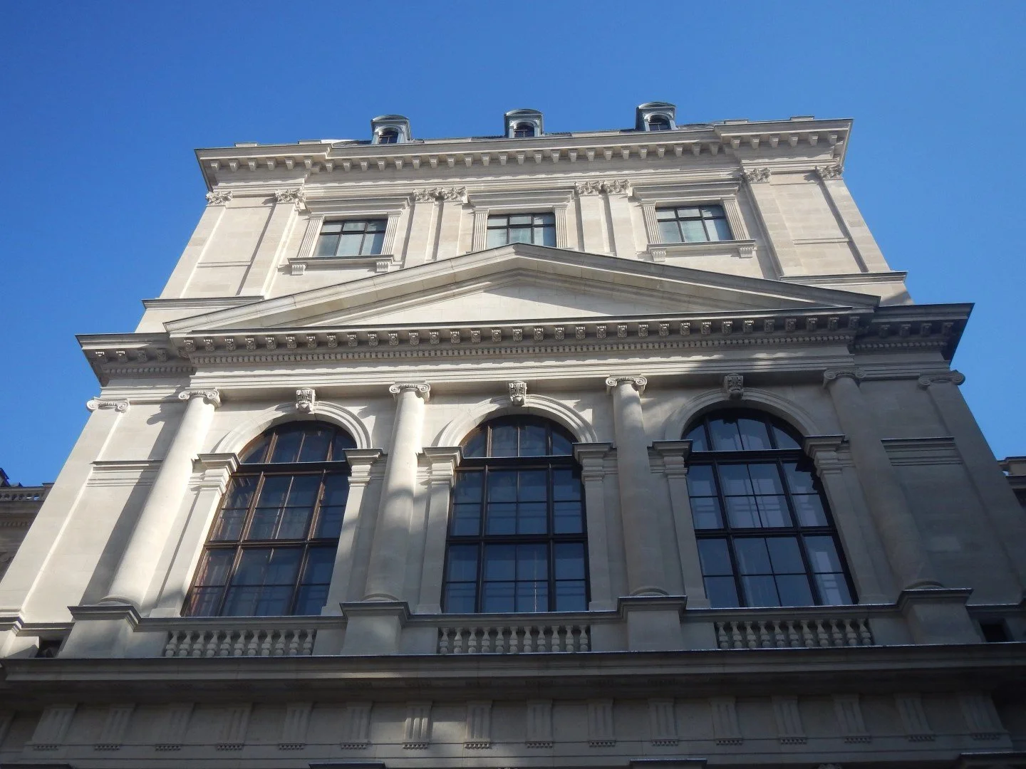 Ein historisches Gebäude mit barocker Architektur, großen Fenstern und klassischen Säulen, fotografiert von unten gegen den blauen Himmel.