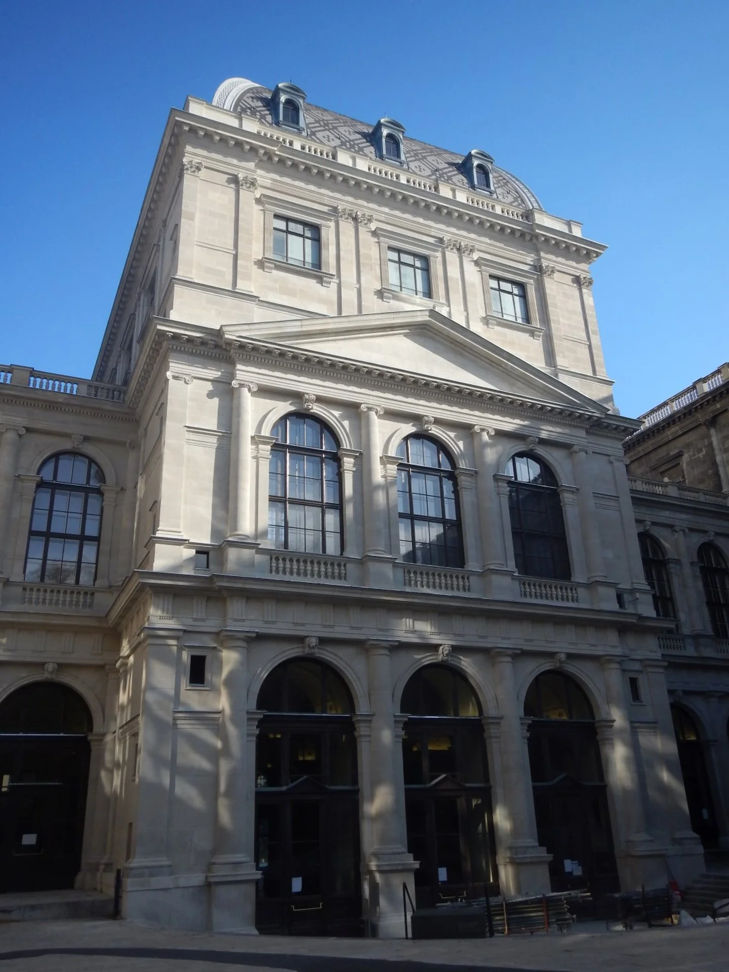 Ein historisches, dreistöckiges Gebäude mit großen Fenstern im klassizistischen Stil, auf einem Platz vor einem klaren blauen Himmel.