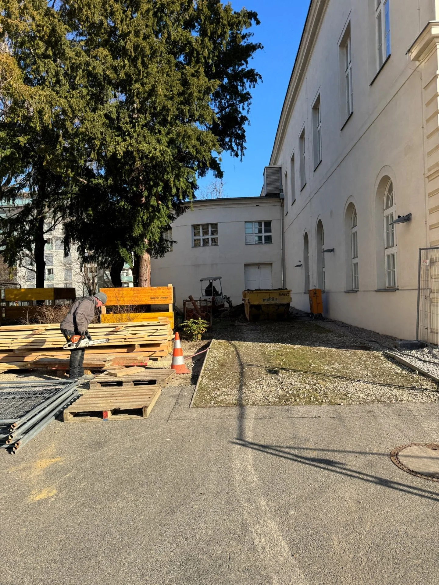 Bauarbeiten im Außenbereich eines Gebäudes mit einem Arbeiter, der Holz verarbeitet, Kisten, Verkehrshütchen und Baugeräte, während im Hintergrund Bäume und ein weißes Gebäude zu sehen sind.