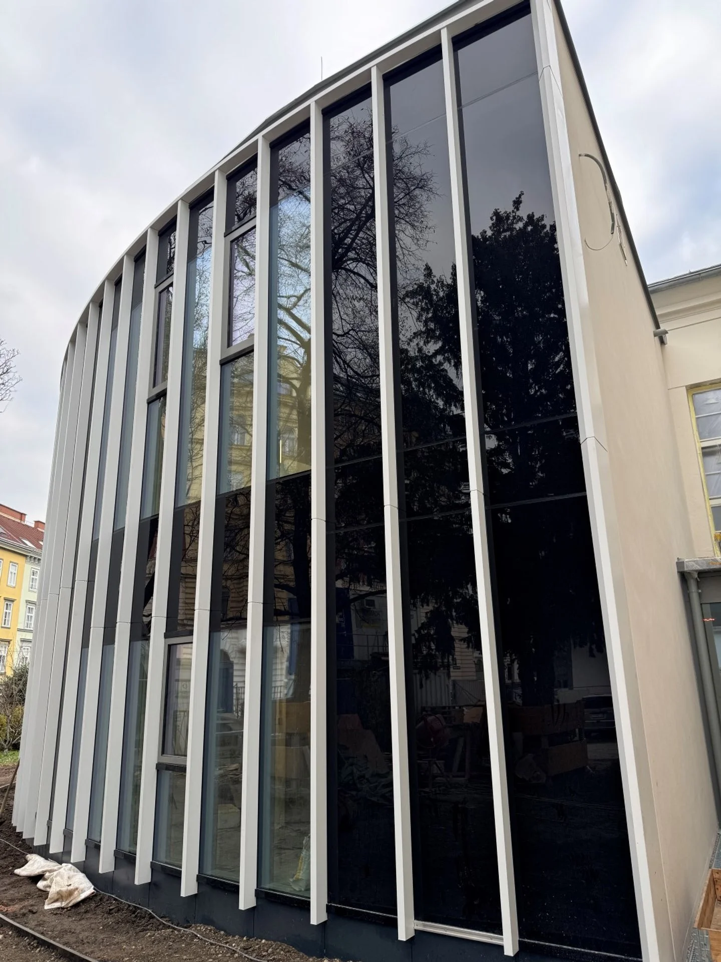 Modernes Gebäude mit großen dunklen Glasfenstern und weißen vertikalen Lamellen an einer Ecke, umgeben von einem Gehweg und umliegenden Gebäuden.